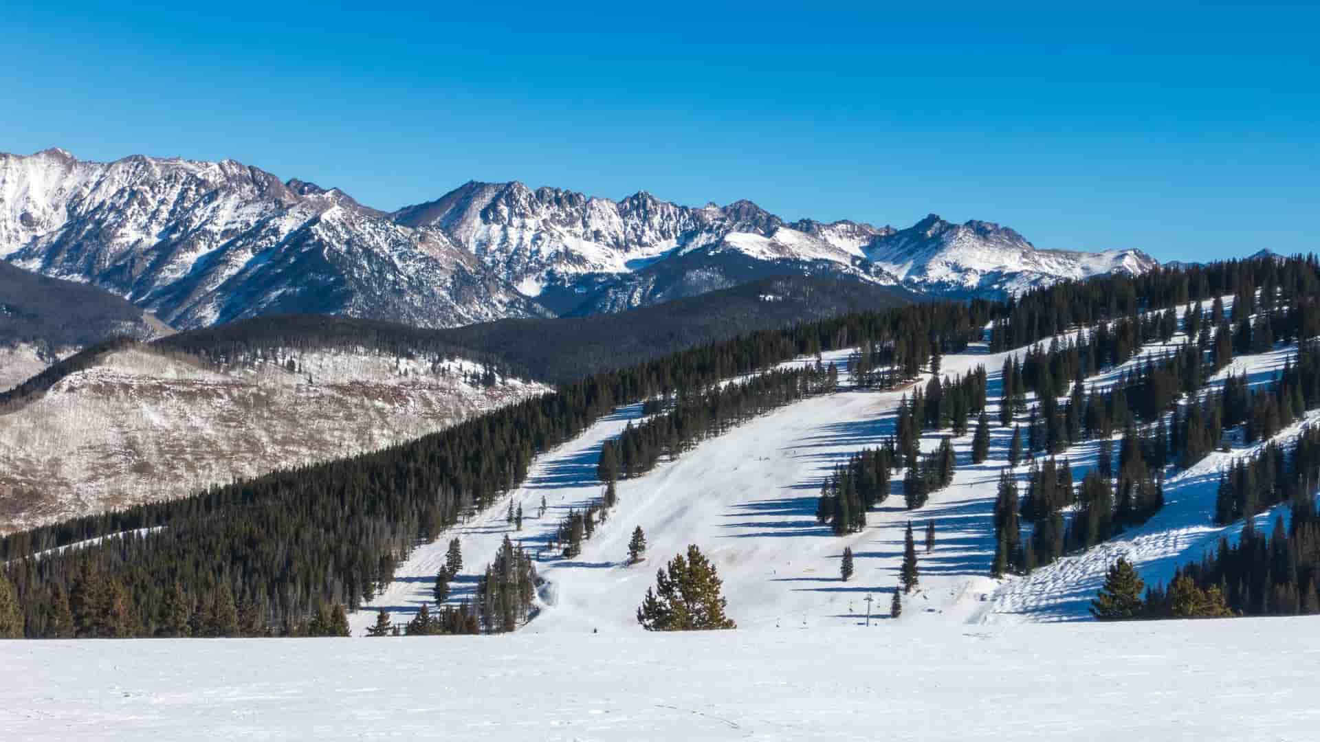 A panoramic view of the Keystone Ski Resort in Colorado, USA, featuring a wide, snow-covered ski run descending through a dense dark evergreen forest, set against a backdrop of rugged, snow-capped Rocky Mountains under a clear blue sky.