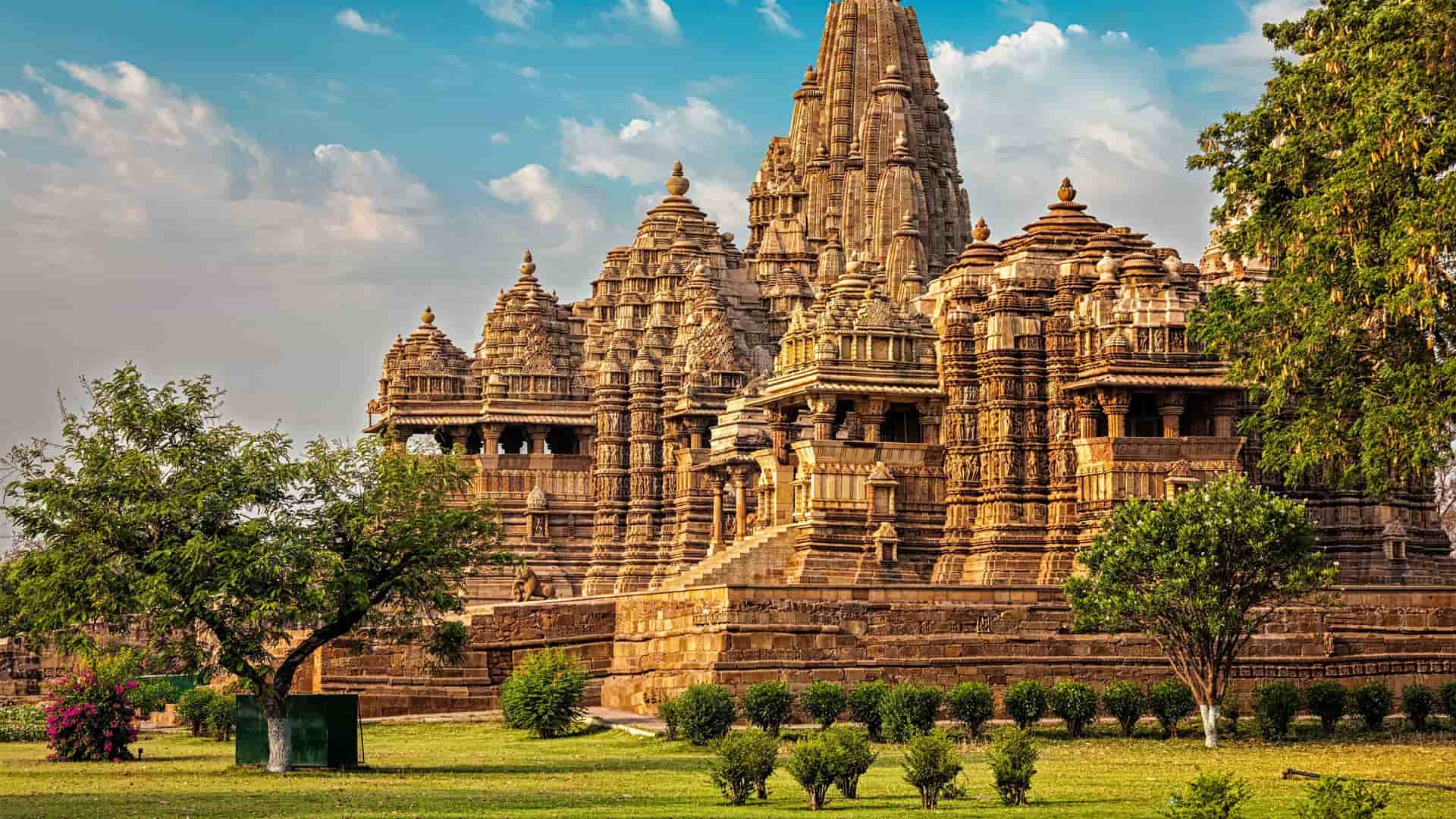 "A magnificent view of the Kandariya Mahadeva Temple, the largest of the ancient Hindu temples in Khajuraho, India, with its intricately carved spires and detailed stone architecture, surrounded by green lawns and trees.  "