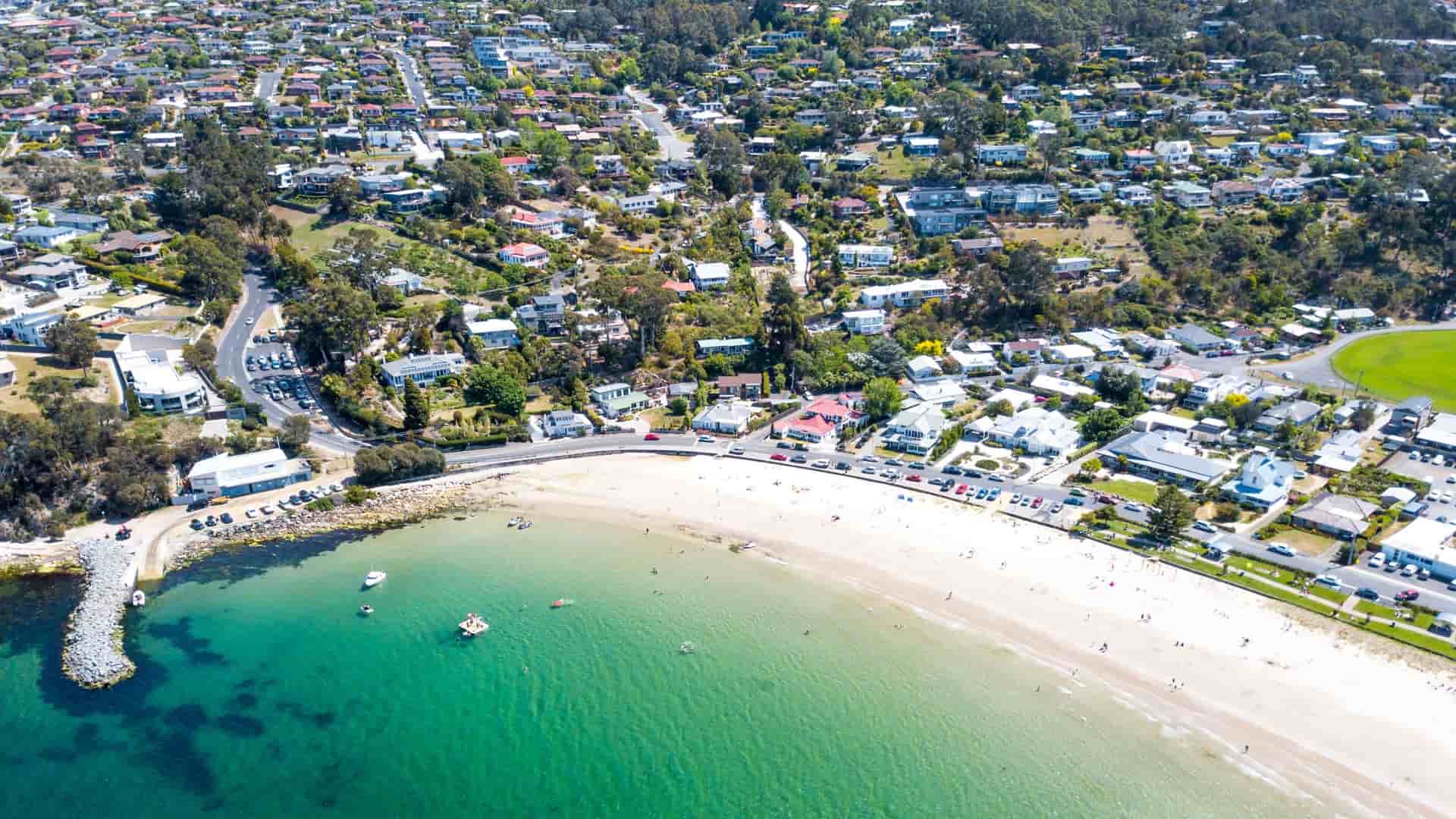 An aerial view of Kingston Beach, a residential suburb near Hobart, Tasmania, Australia. The image shows the curved white-sand beach and the adjacent neighborhood of houses and streets, bordering the calm, blue waters of the River Derwent estuary.