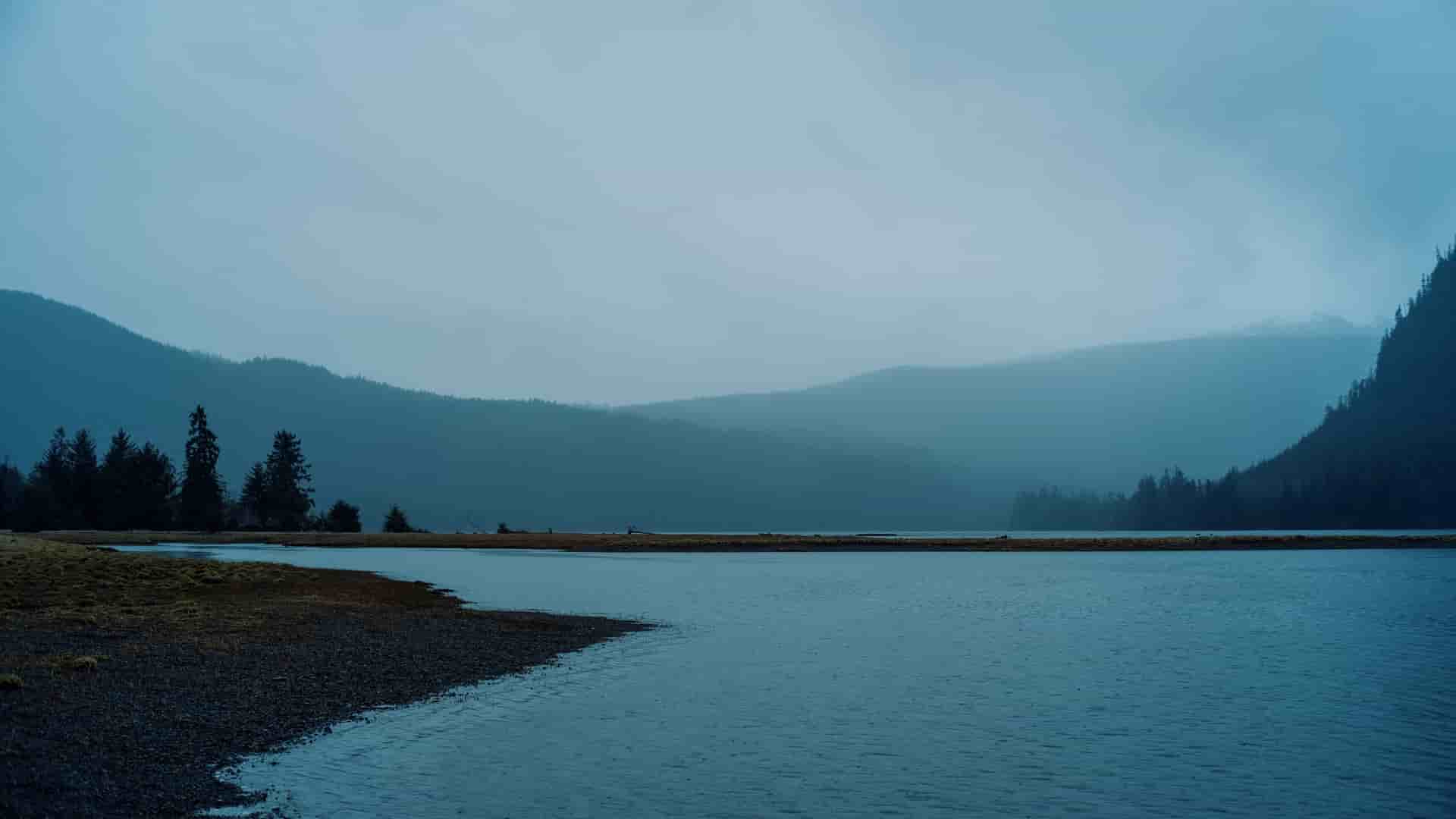 A tranquil, moody landscape of Klawock, Alaska, with a calm lake reflecting dark, forested mountains in the distance under an overcast and hazy sky.
