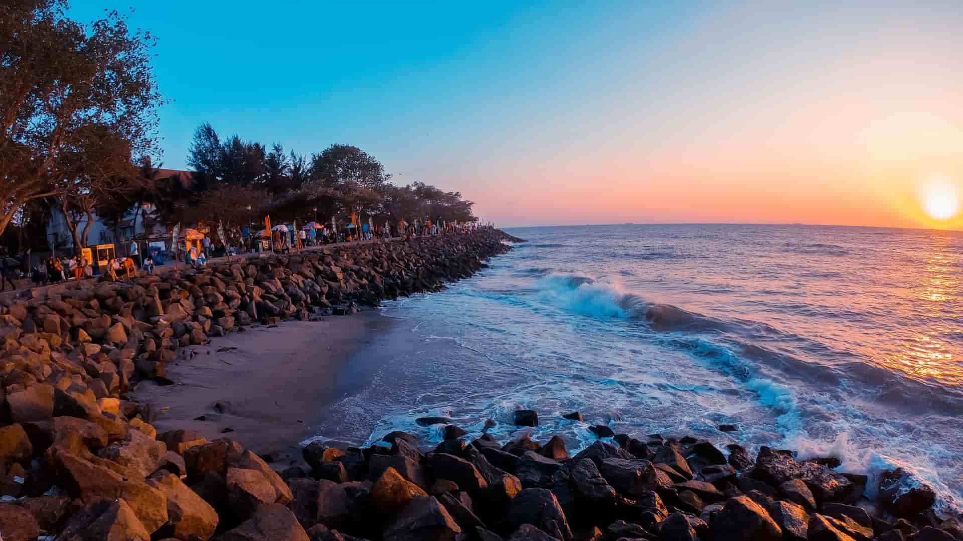 A vibrant sunset view over the ocean in Kochi (Cochin), India, with waves crashing against a rocky coastline and a lively walkway of people and trees.