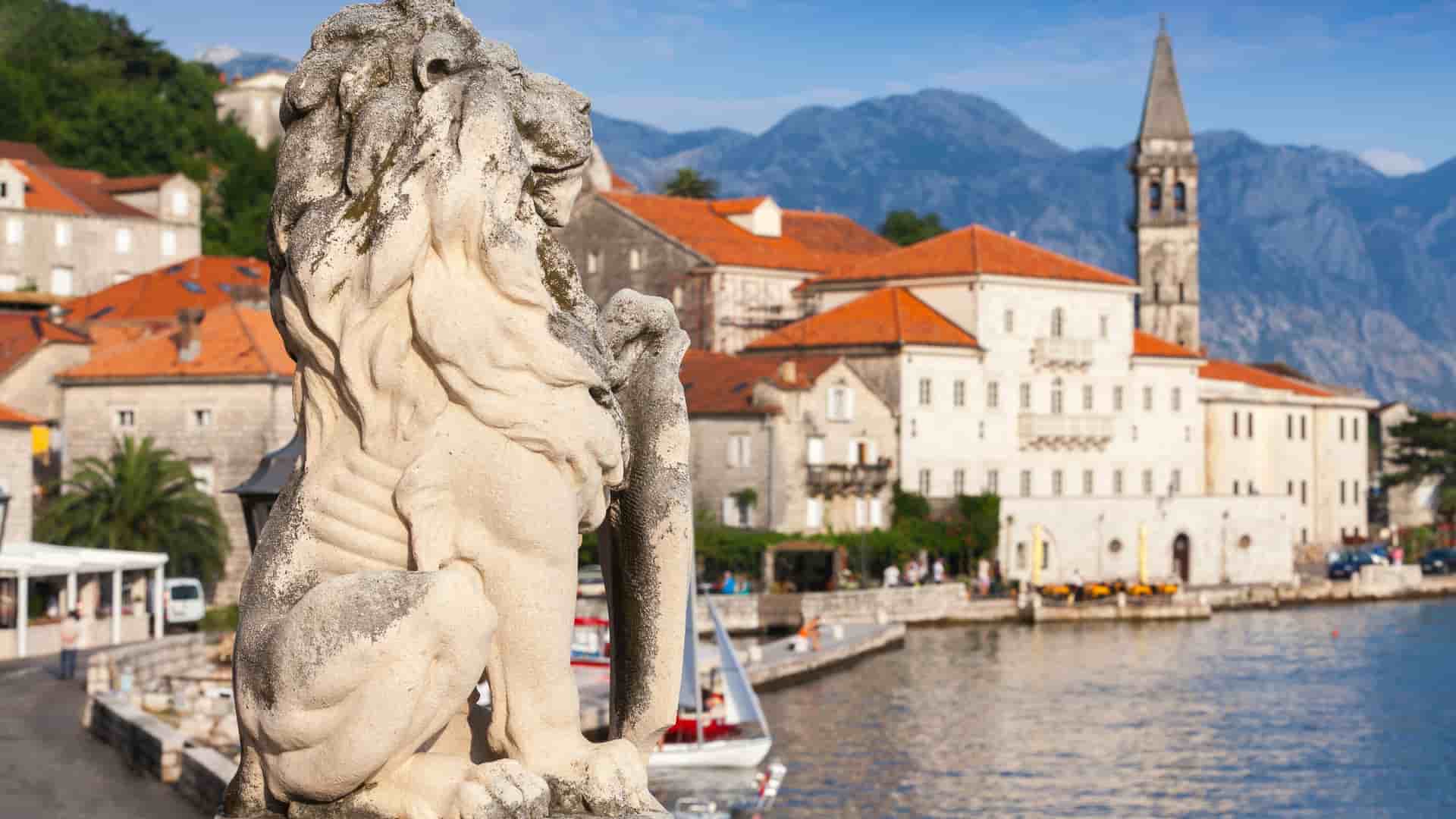A stone lion statue in the foreground with the beautiful city of Perast and the Bay of Kotor in Montenegro in the background, featuring red-roofed buildings and a church steeple by the water.