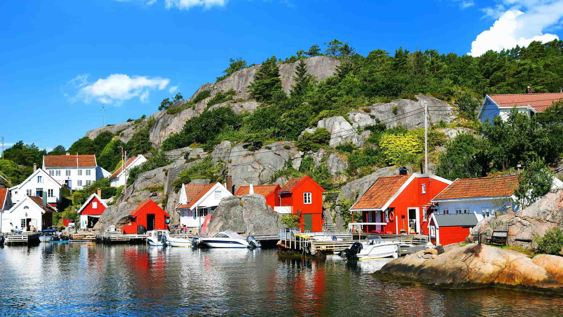 A beautiful Norwegian landscape with traditional red and white wooden houses on the water's edge, nestled against a rocky, forested hillside under a clear blue sky.