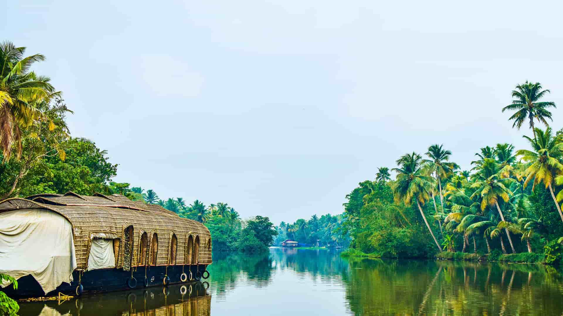"A scenic view of a traditional houseboat on the tranquil backwaters of Kumarakom, Kerala, India, with lush palm trees and tropical vegetation lining the banks.  "