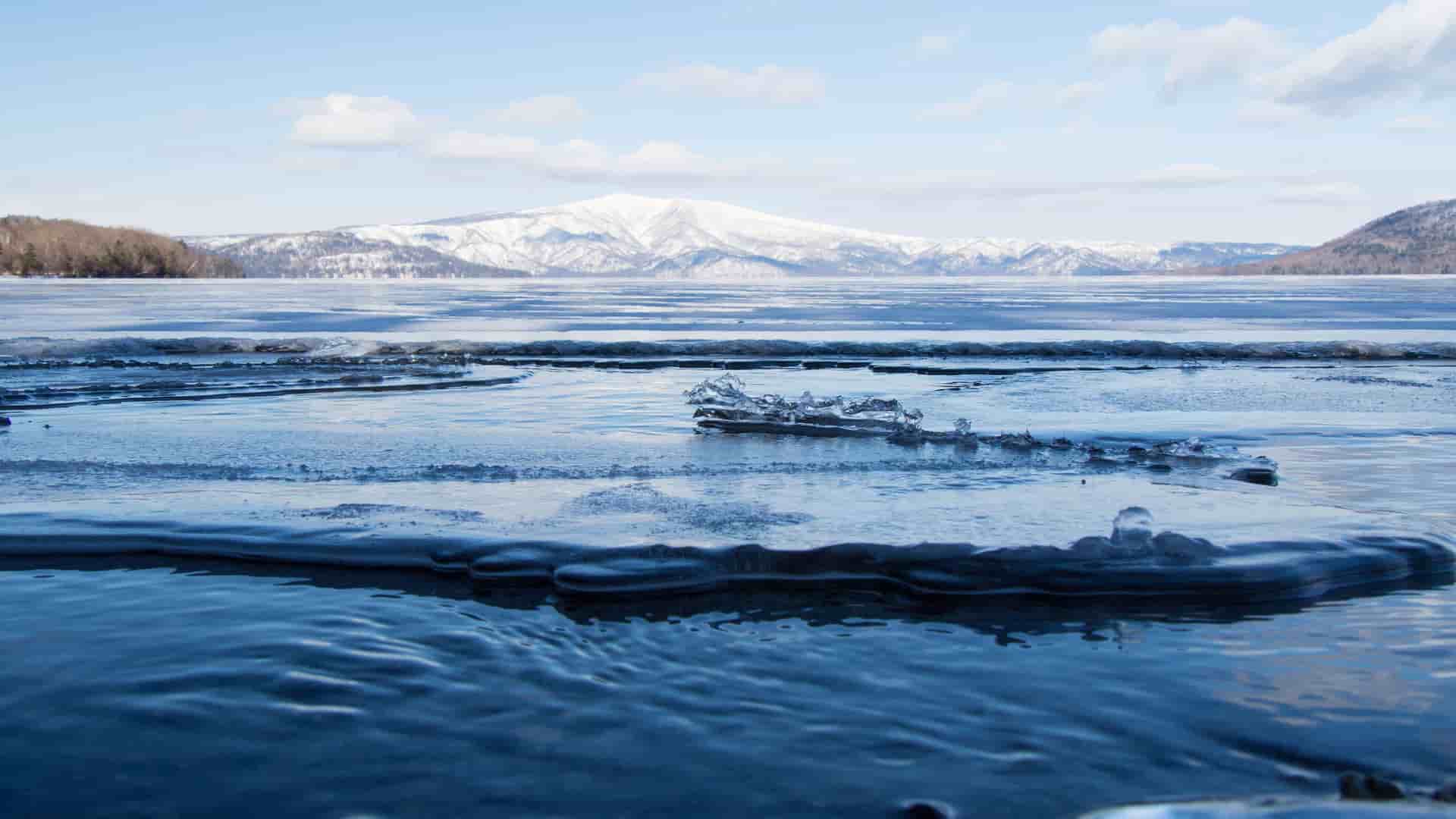 A scenic winter view of Lake Akan in Kushiro, Japan, with a layer of thin ice on the water in the foreground and snow-covered mountains visible in the distance under a blue sky.