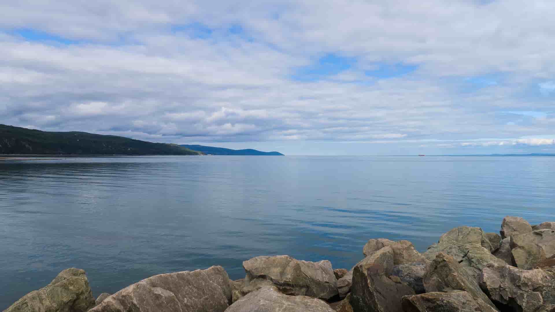 "A tranquil scenic shot of the St. Lawrence River in La Malbaie, Quebec, Canada, showing the calm blue water stretching to the horizon, with rocky shores and green hills under a cloudy sky.  "