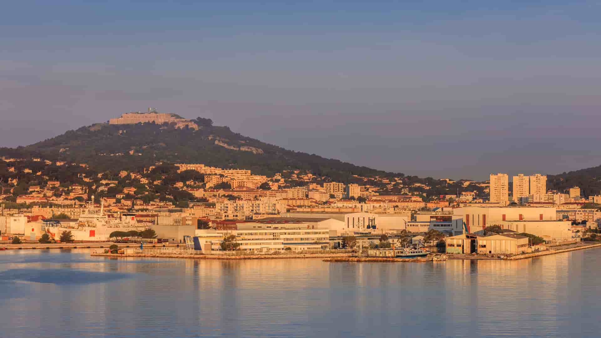 A serene sunrise over the harbor of La Seyne-sur-Mer, with a bustling shipyard in the foreground and a historic fort perched atop a lush green hill.