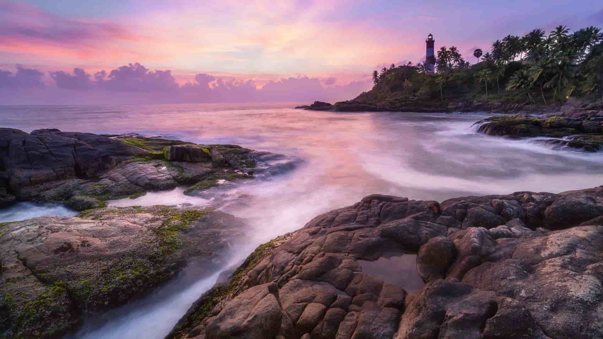 A dramatic sunset over the Laccadive Sea, with a tall lighthouse on a tropical, palm-fringed cliffside and waves crashing against a rocky shoreline.