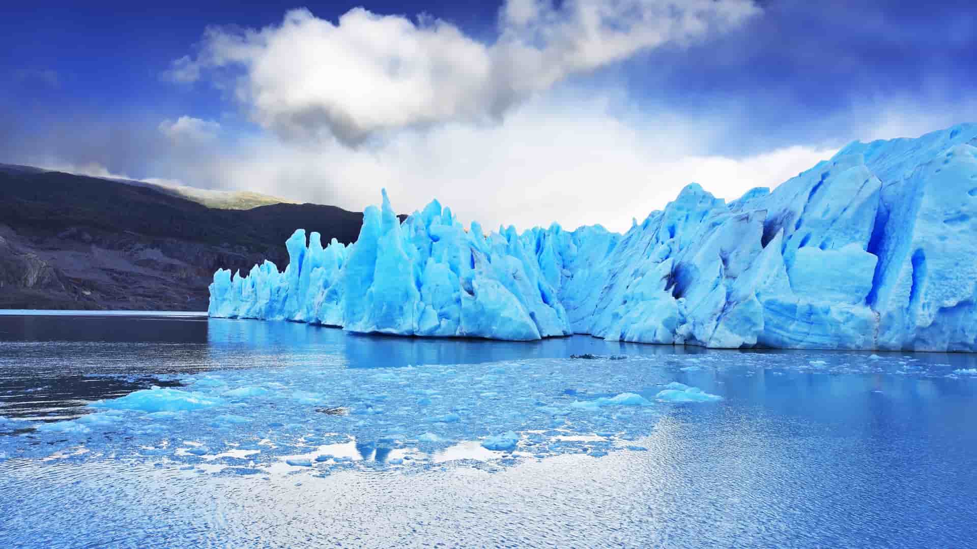 A large, stunningly blue iceberg in Laguna San Rafael, with smaller chunks of ice floating in the water and a rugged mountain in the background.