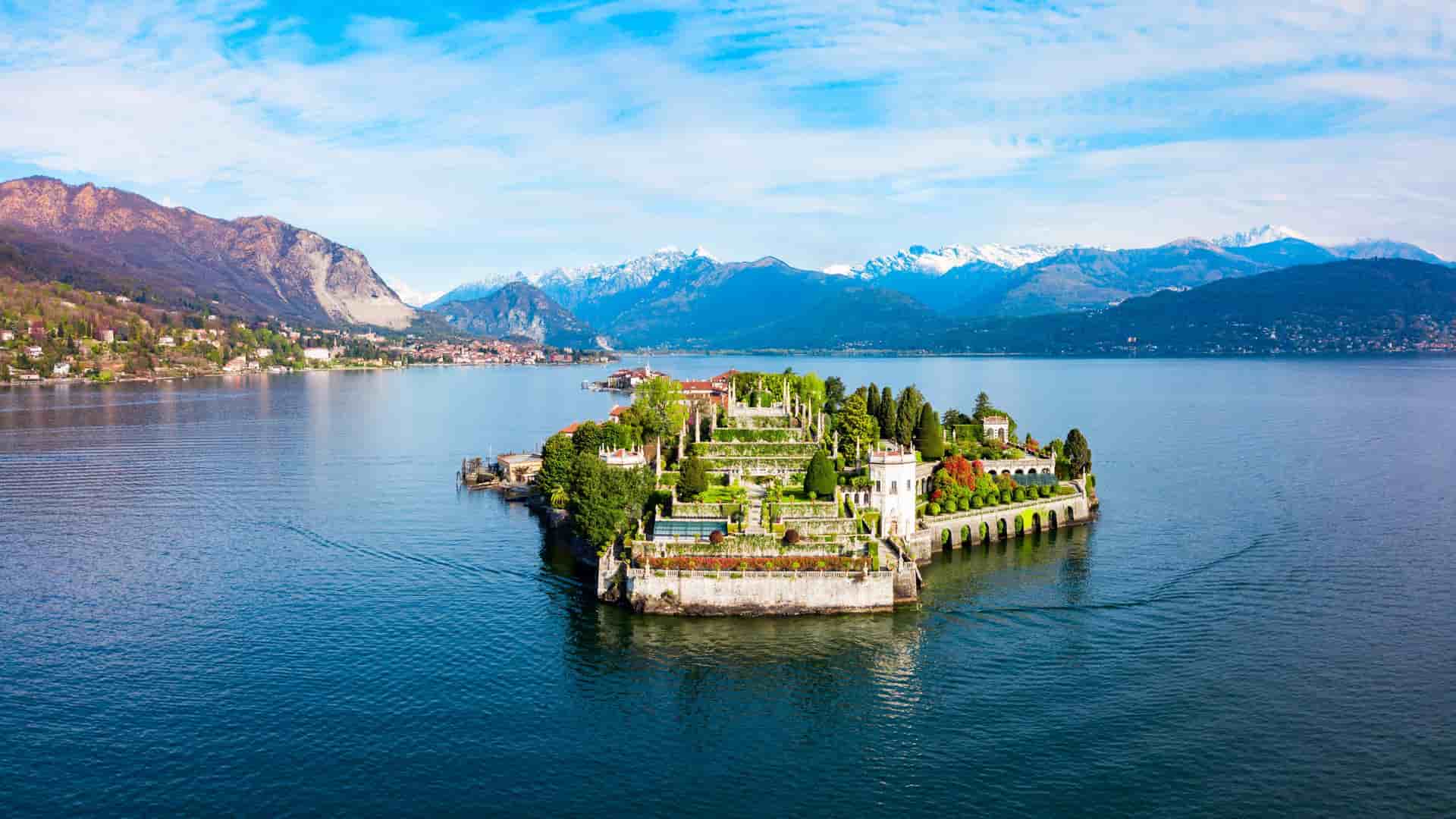 "An aerial view of the beautiful Isola Bella island on Lake Maggiore, Italy, showing the elaborate Baroque Borromeo Palace, its terraced gardens, and the snow-capped Swiss Alps in the background. "