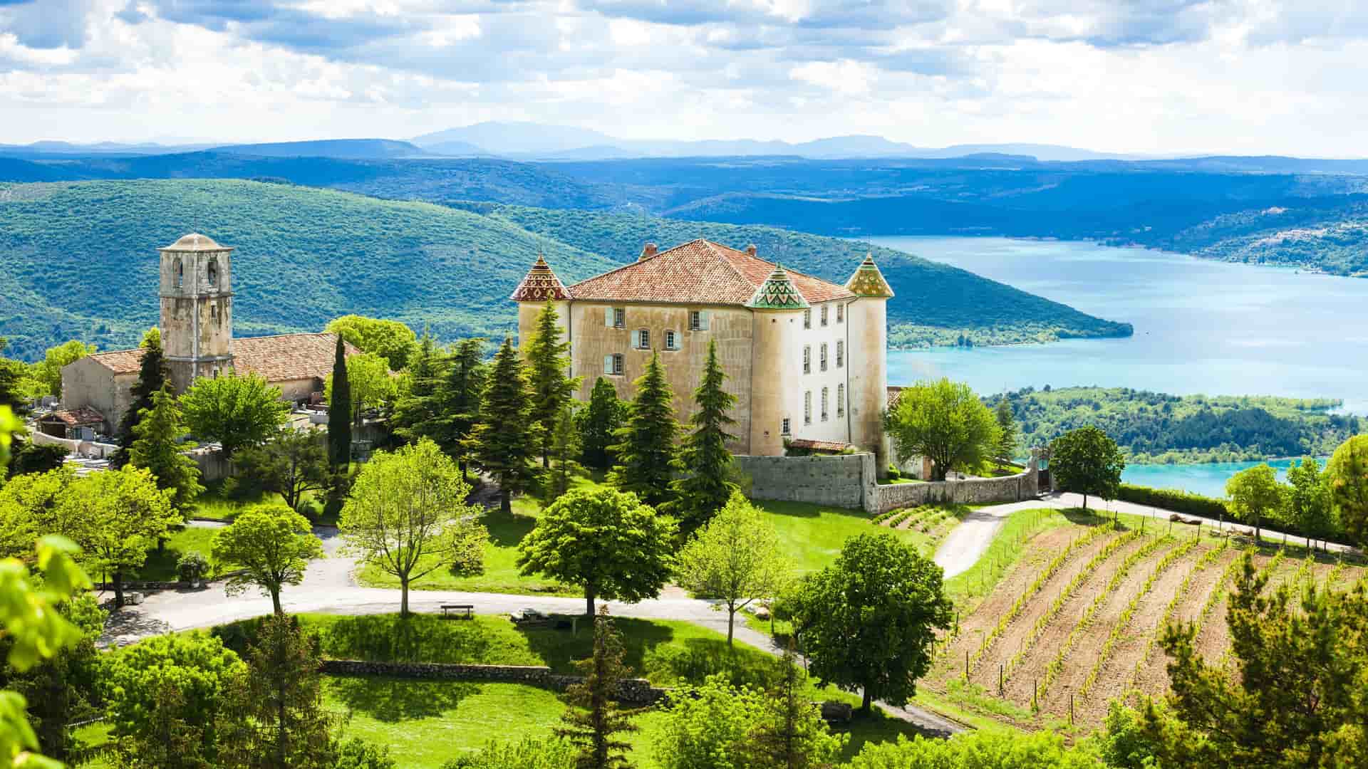 A panoramic view of the historic Chateau de Aiguines perched on a hill overlooking the turquoise waters of the Gorges du Verdon in Provence, France, with lush green hills and a vineyard in the foreground.