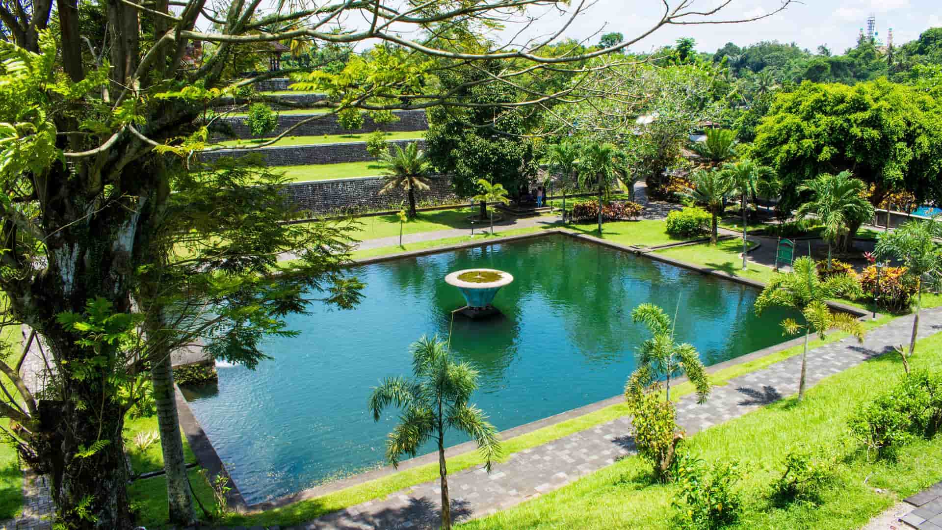 A lush, tropical garden with a rectangular man-made pond and a fountain in the center, surrounded by trees and walking paths at the Taman Narmada Park near Lembar, Bali.