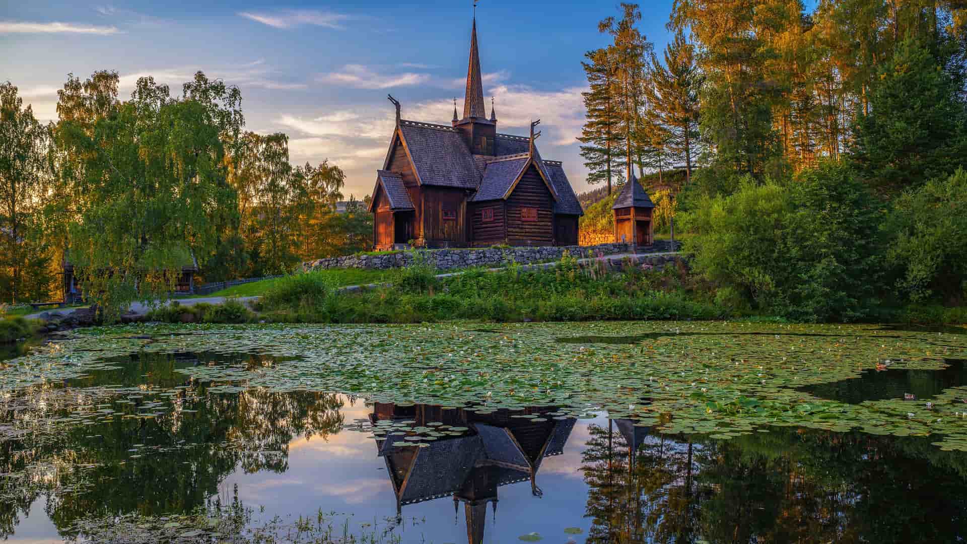 "A serene lake reflecting the historic Garmo Stave Church in Lillehammer, Norway, surrounded by lush green trees and a tranquil natural landscape under a bright evening sky.  "