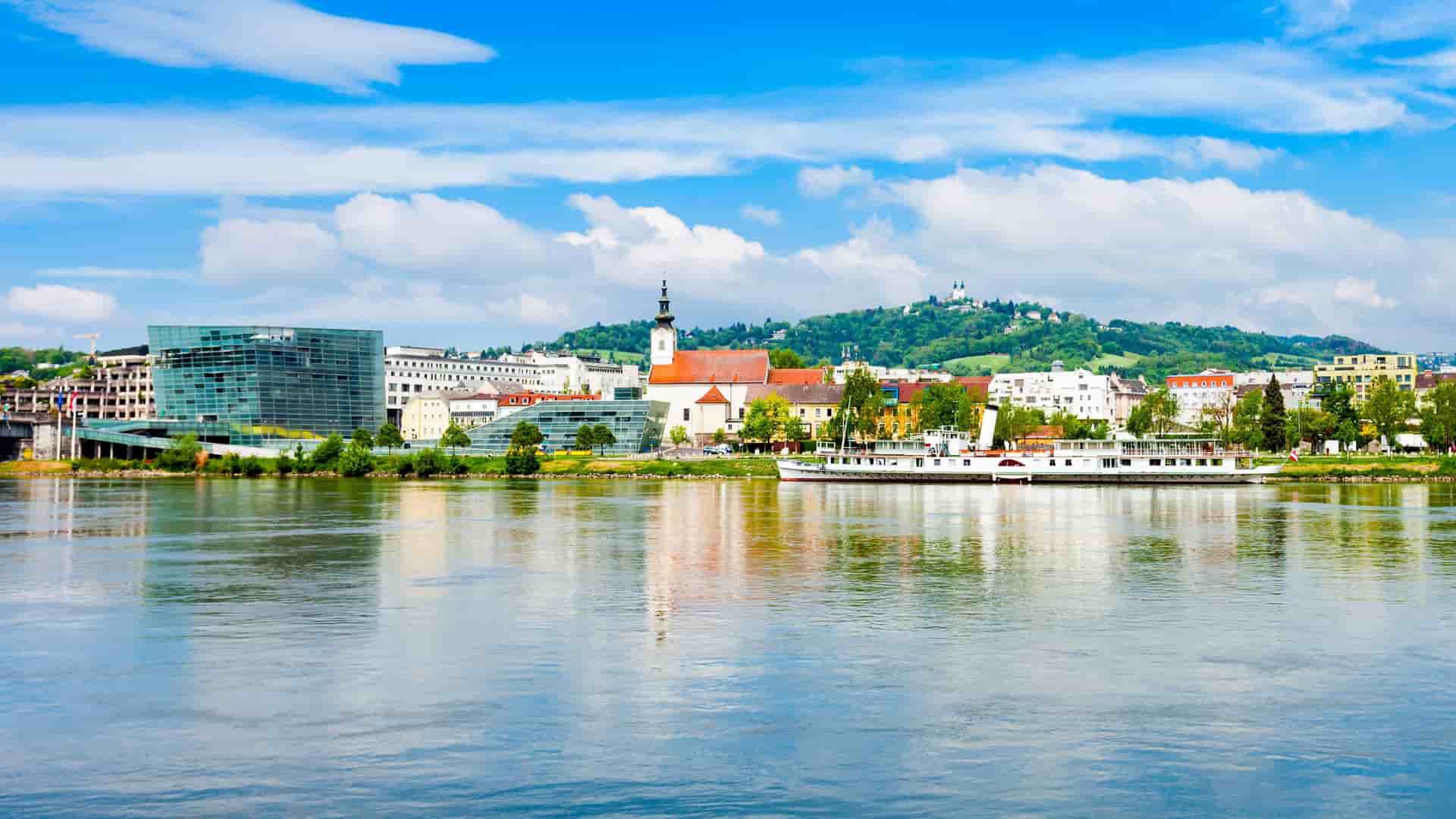 A vibrant panoramic shot of the Linz skyline along the Danube River, showcasing the old town's historic buildings, a modern glass structure, and a boat docked on the water.