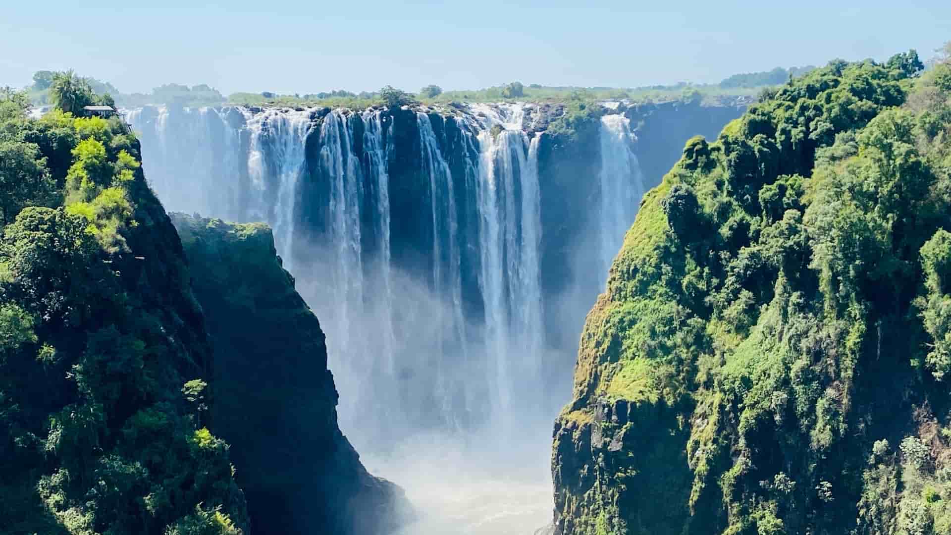 A huge curtain of Victoria Falls water plunges into a gorge surrounded by lush, mist-covered green cliffs near Livingstone, Zambia.