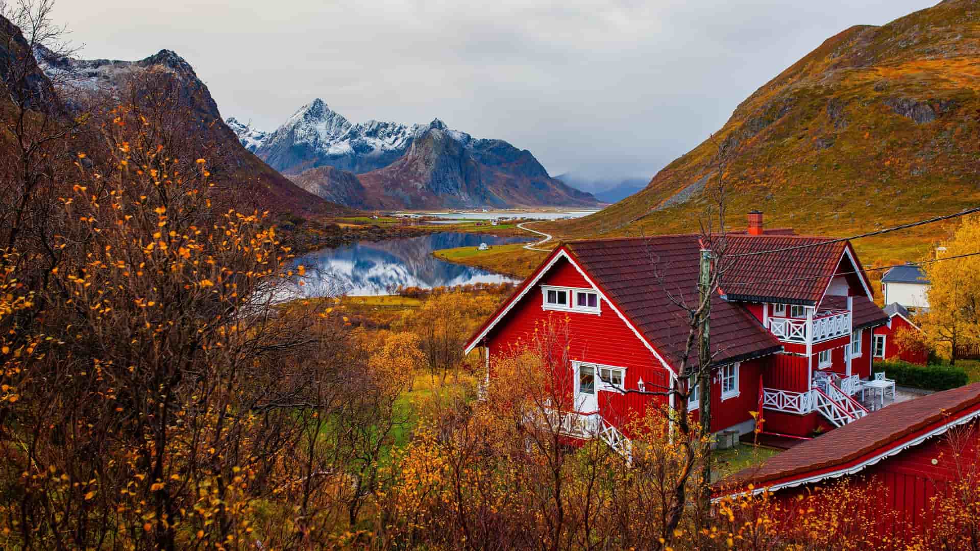 A breathtaking autumn view of a traditional red Norwegian house nestled among golden foliage near Loen, with majestic snow-capped mountains reflected in a calm lake.