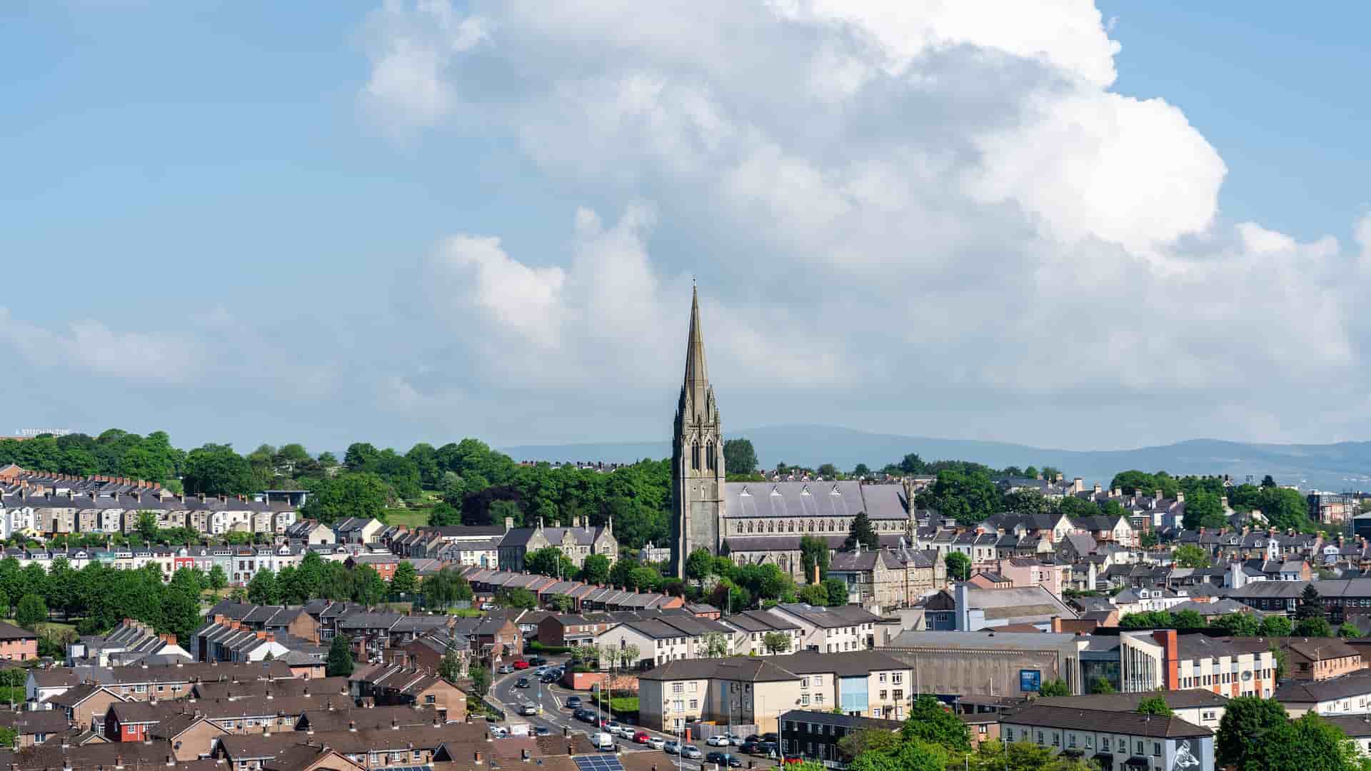 A scenic view of Londonderry, Northern Ireland, with the neo-gothic St. Eugene's Cathedral standing prominently above the cityscape and surrounding green hills.