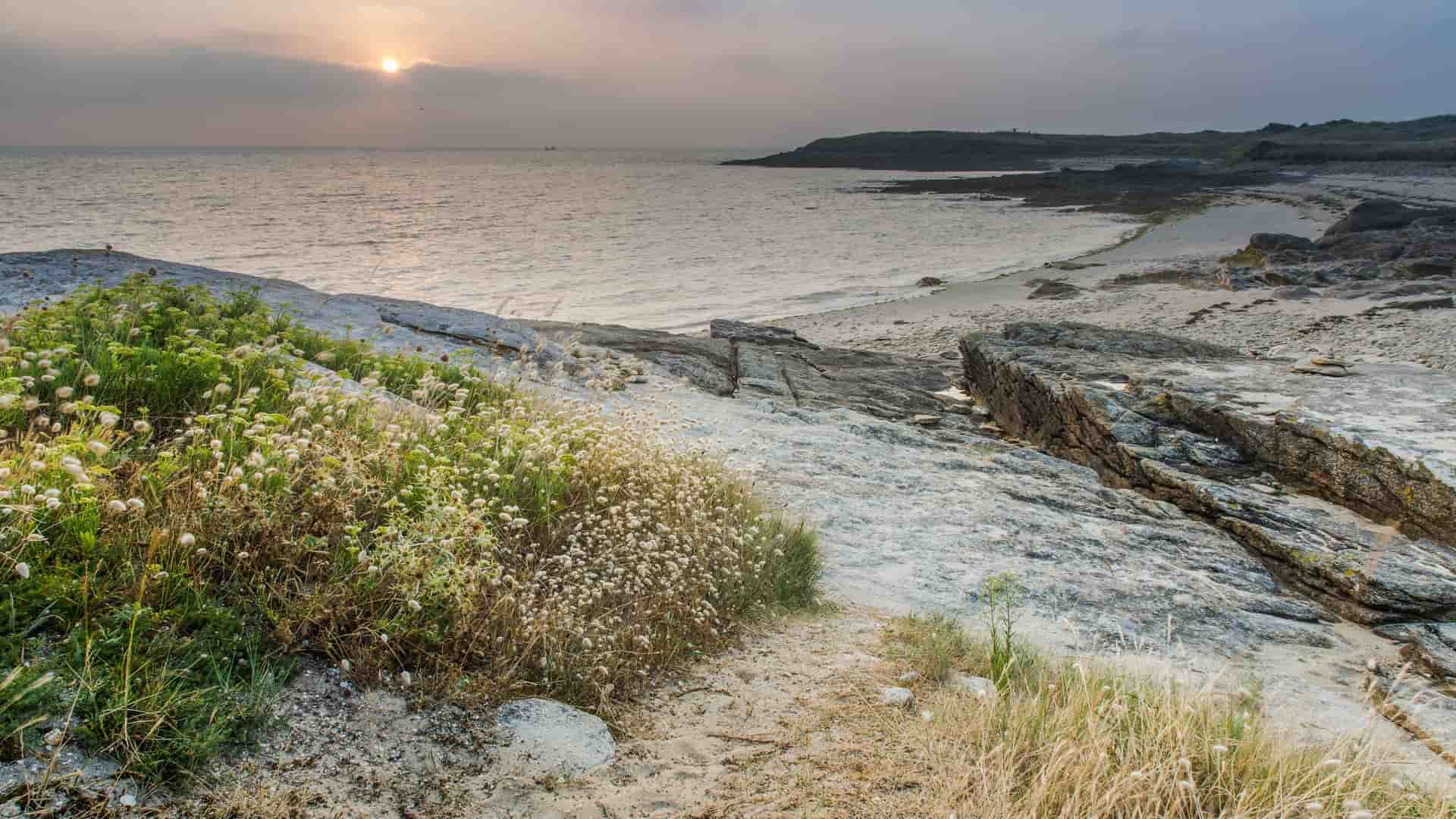 A serene sunset view over the rocky shoreline near Lorient, France, with a tranquil sea and sparse vegetation on a white-pebbled beach under a hazy sky.