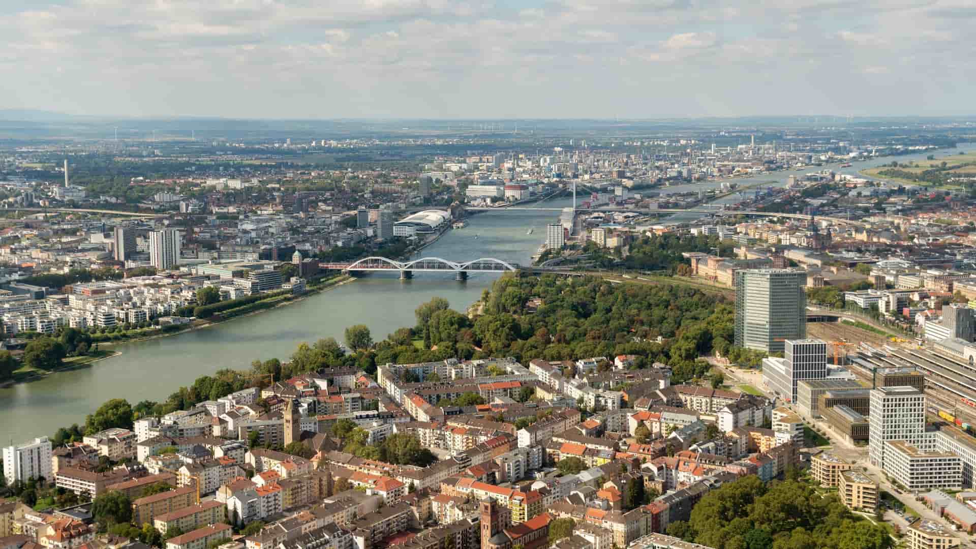 An aerial panoramic shot of Ludwigshafen and Mannheim in Germany, showcasing the Rhine River flowing through the cities, with bridges and modern high-rises.