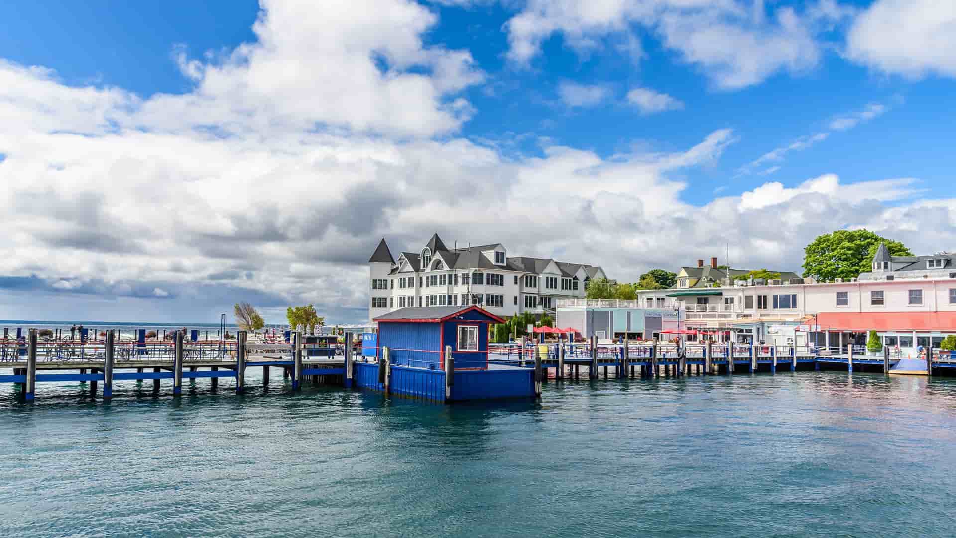 A vibrant shot of the waterfront on Mackinac Island, Michigan, with a colorful blue boathouse and a row of classic white and red buildings under a bright blue sky with puffy white clouds.