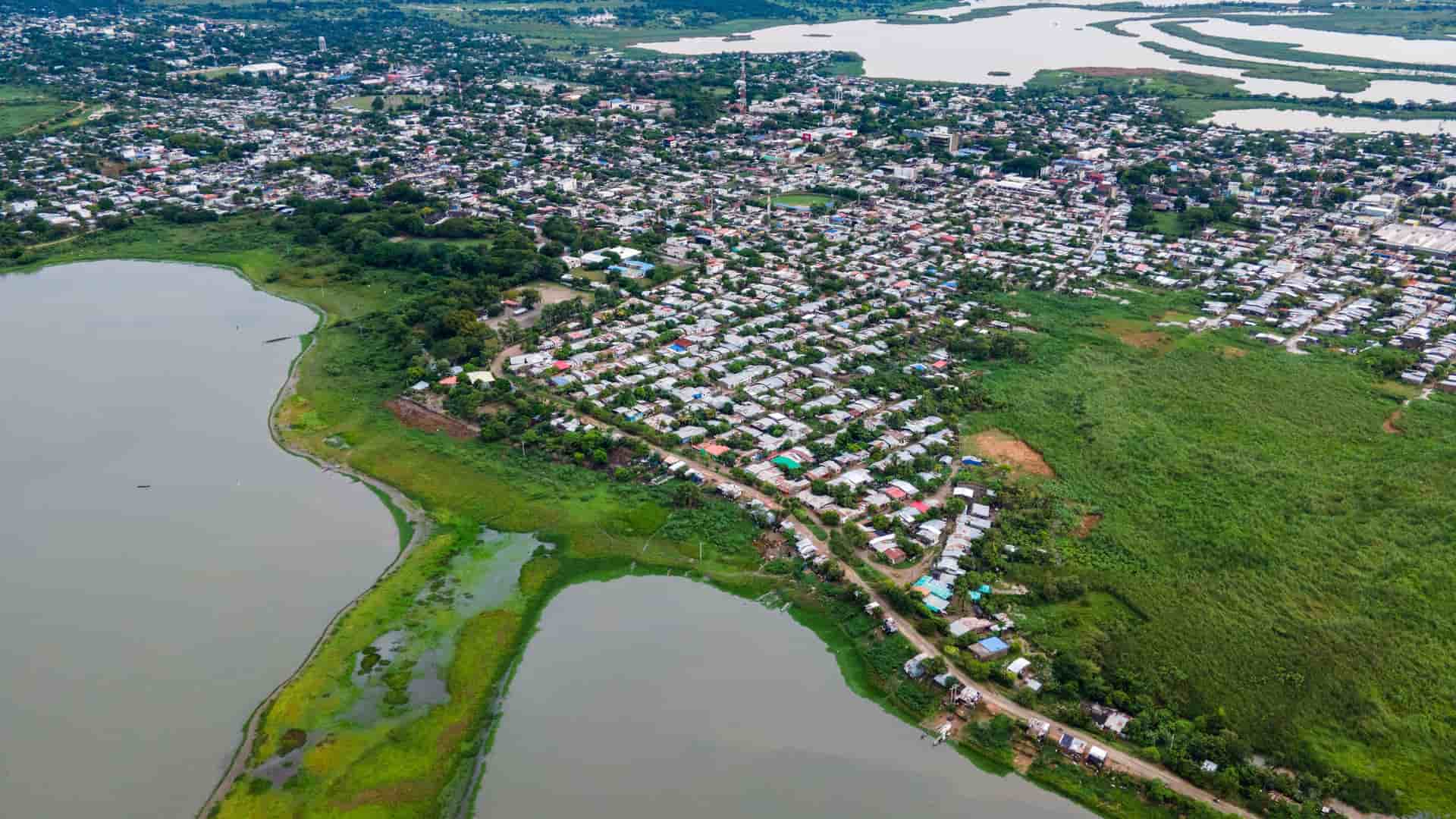 Aerial view of Magangué, Colombia, a densely built town with colorful roofs, bordered by a murky river/lake and lush green land, under an overcast sky.