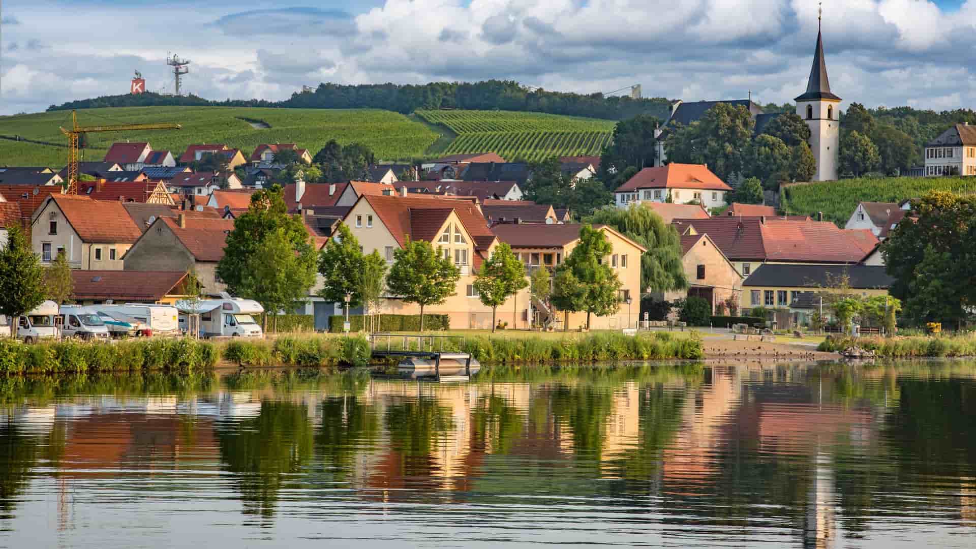 A serene view of a small German village on the Main-Danube Canal, with buildings and a church spire reflecting in the calm water.