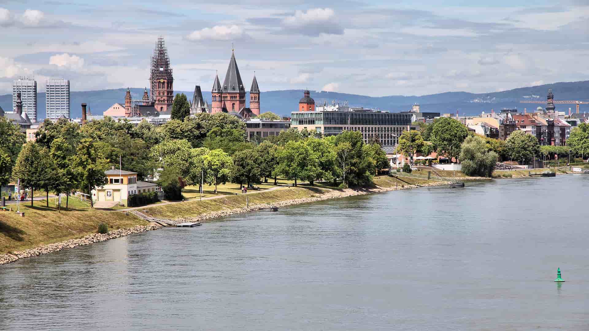 A cityscape of Mainz, Germany, from across the Rhine River, showcasing historic buildings, the distinctive cathedral, and green parkland along the waterfront.