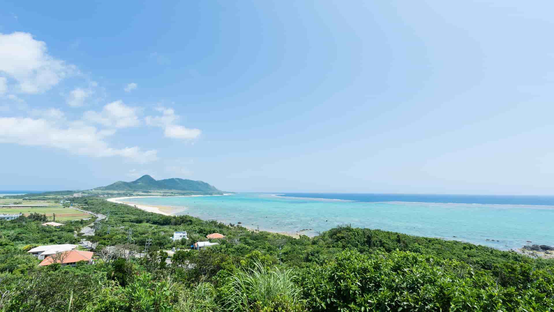 A stunning aerial shot of the coastline in Maizuru, Japan, with turquoise water, a sandy beach, and a lush green landscape under a clear blue sky.