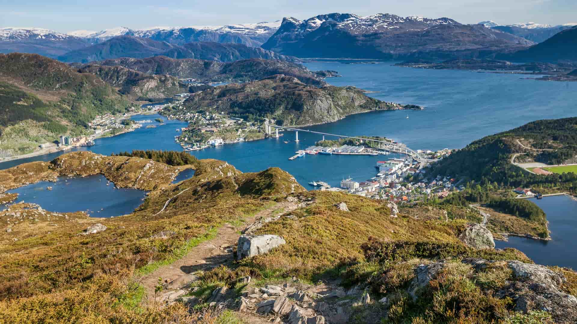 A breathtaking panoramic shot of Måløy, Norway, from a mountain trail, with the town, its bridge, and the surrounding fjord and snow-capped mountains.
