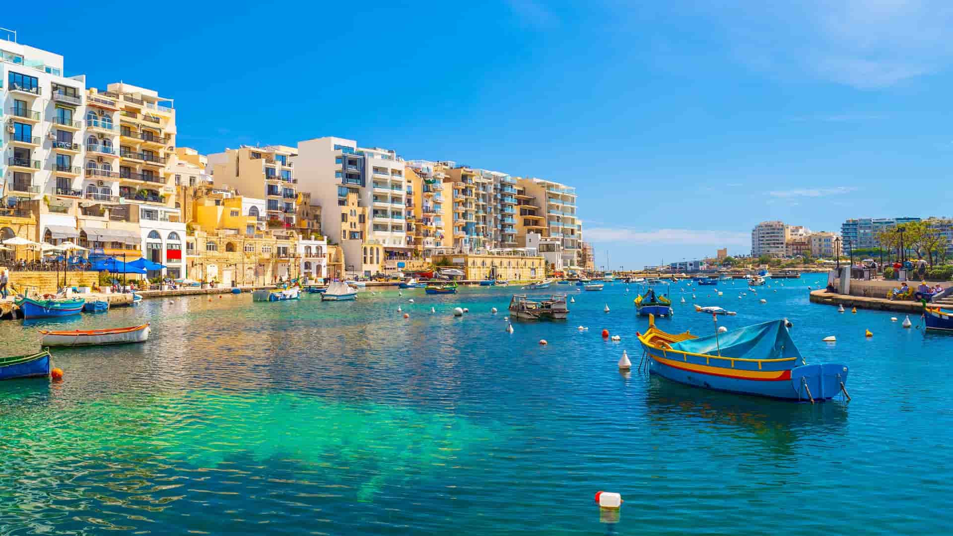 Colorful boats float in the bright turquoise water of a harbor in Malta, backed by white and yellow multi-story waterfront buildings.