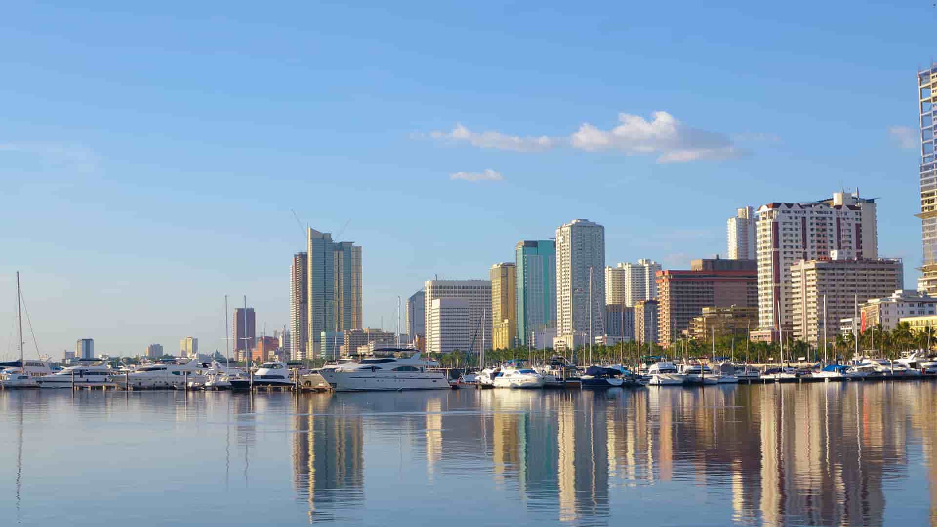 A picturesque view of the modern skyline of Manila, Philippines, with high-rise buildings reflecting on the calm waters of the bay filled with docked yachts and boats.