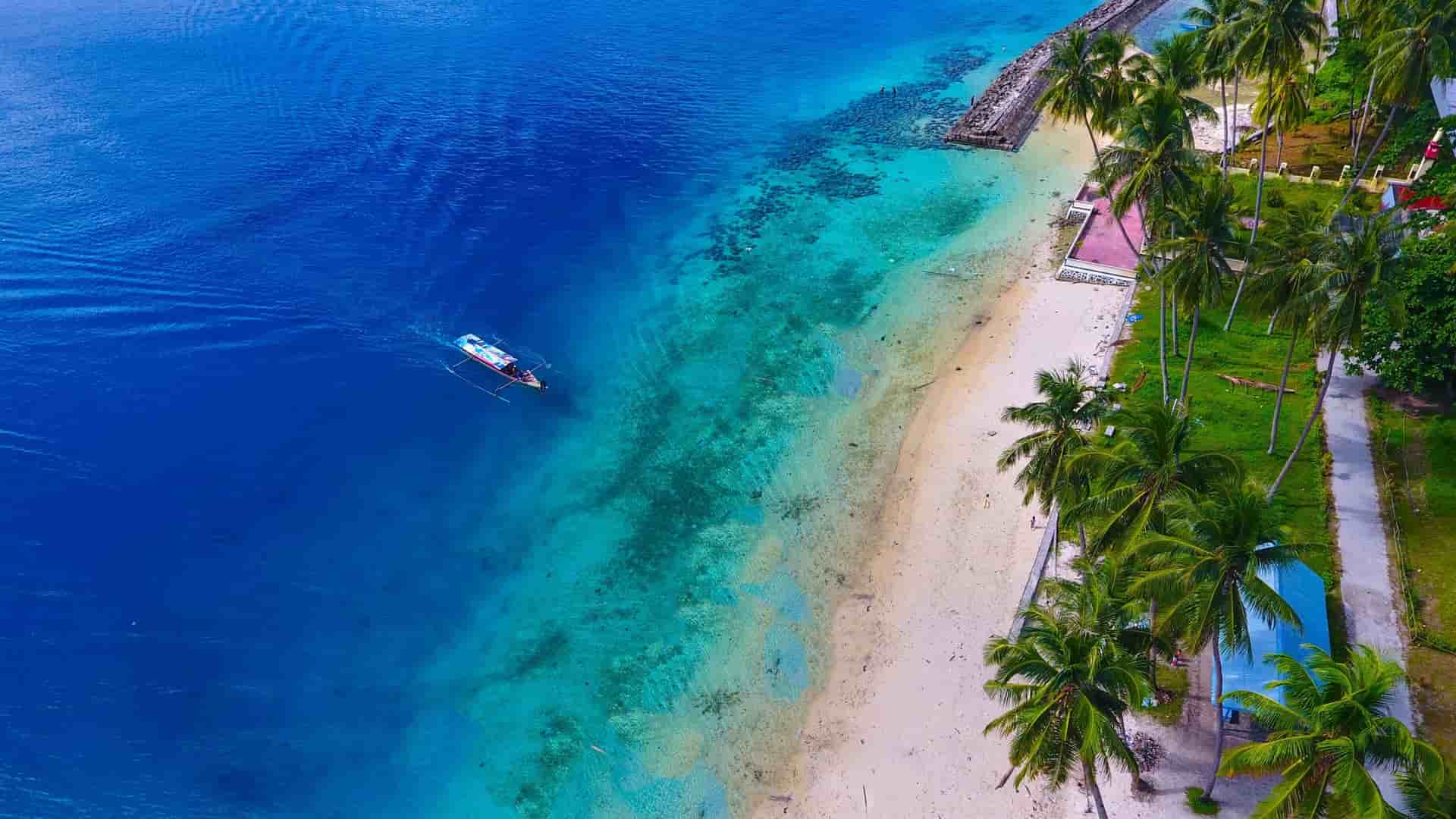 An aerial view of a vibrant blue tropical shoreline in Manokwari, Indonesia, with a small boat in the water, a white sand beach, and palm trees.