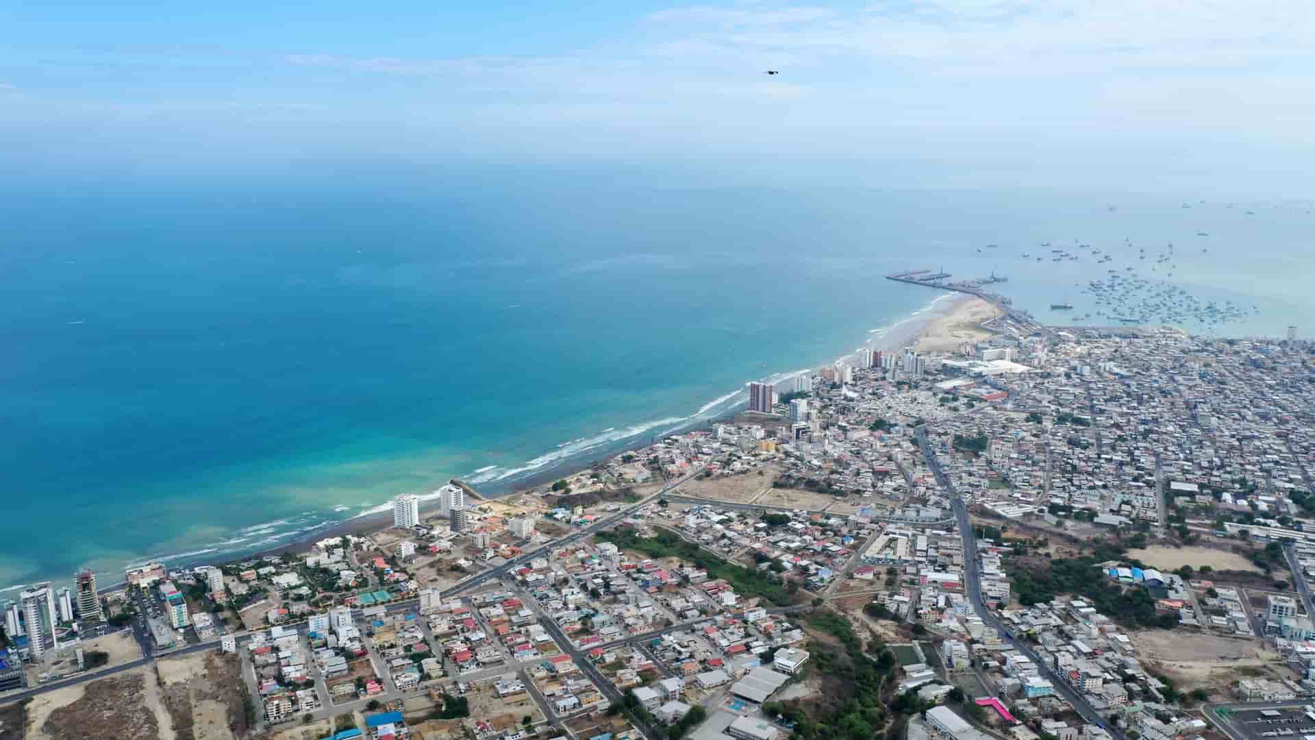 A high-altitude aerial view of a sprawling coastal city with dense housing along a turquoise ocean and a busy harbor filled with boats.