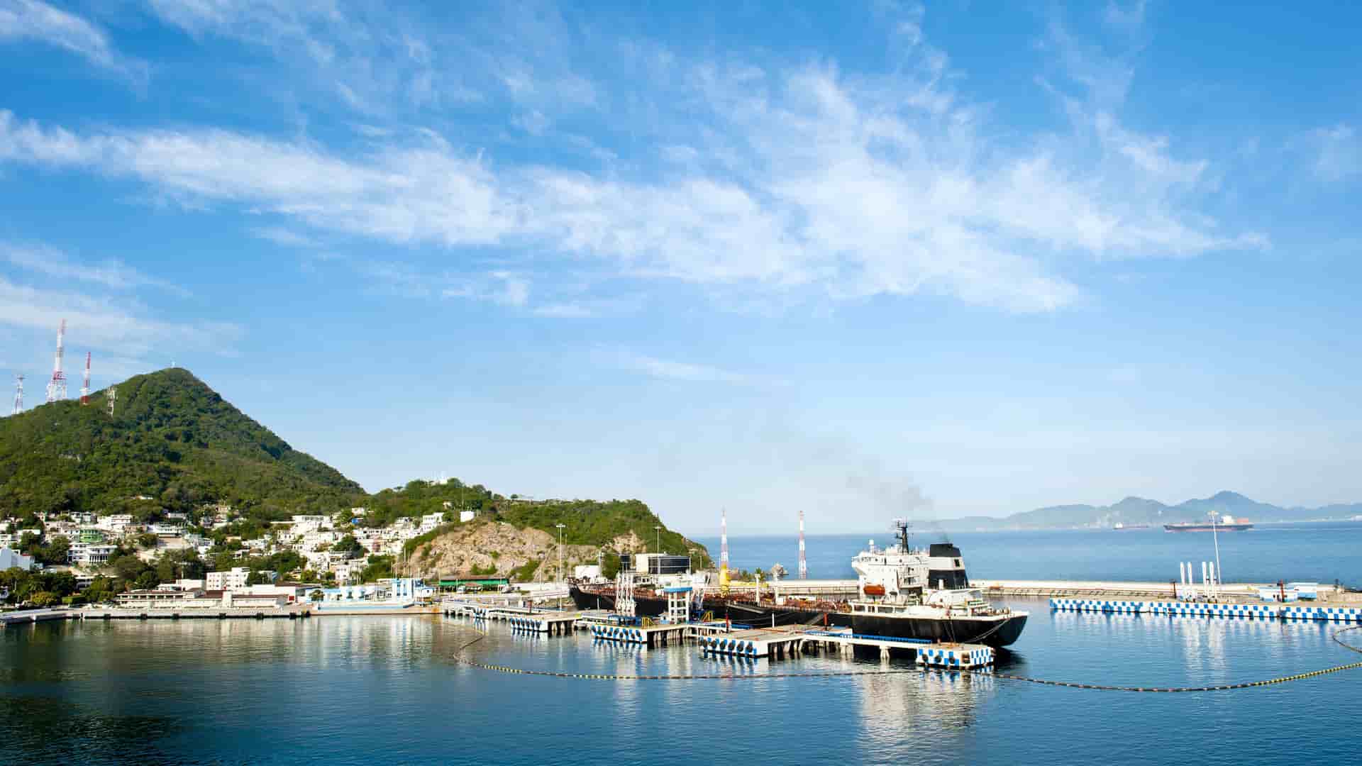 A panoramic view of the bustling harbor of Manzanillo, Mexico, with a large tanker docked and mountains in the background under a bright blue sky.