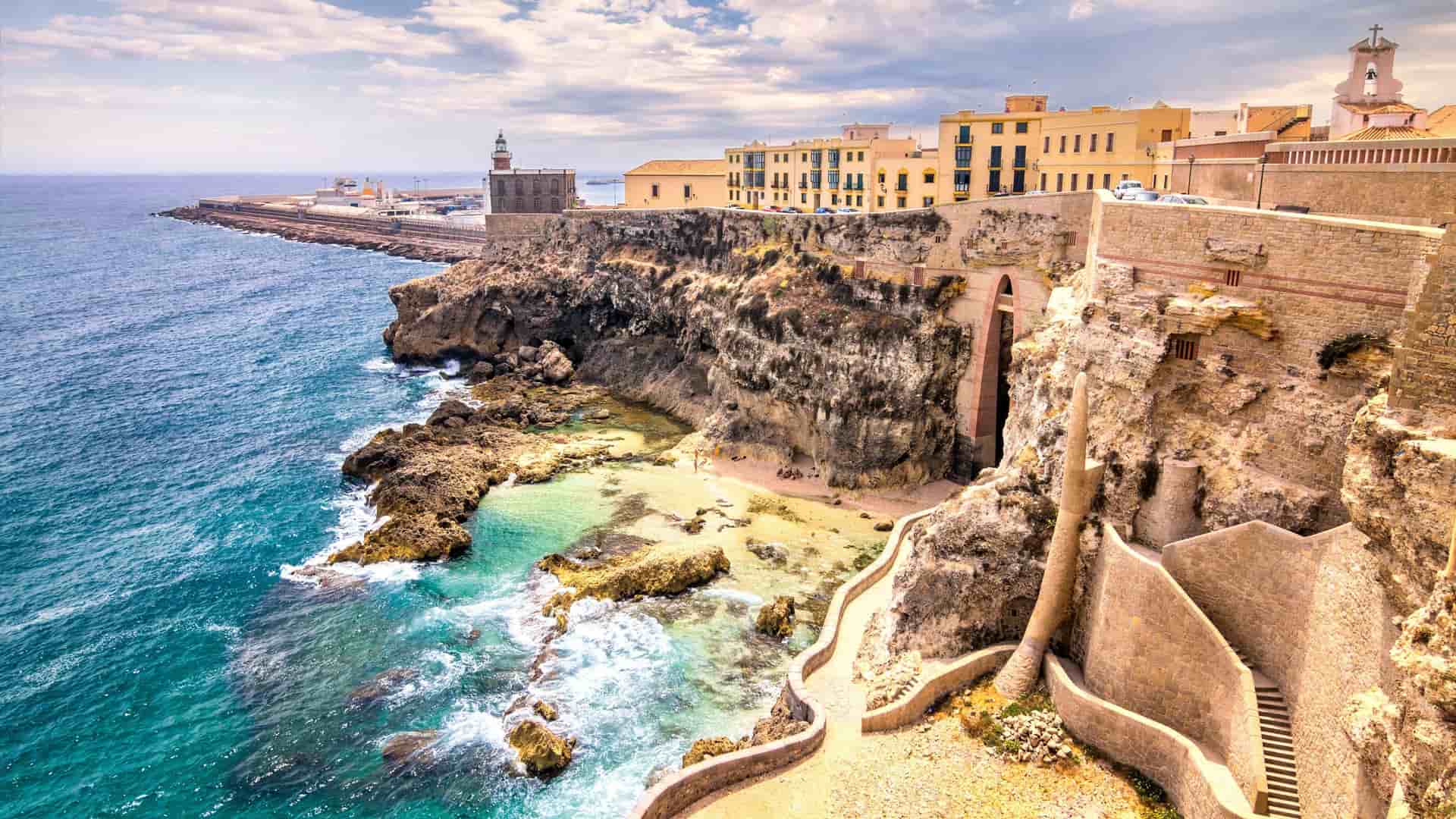 A panoramic view of the fortress walls and cliffs of Melilla, a Spanish enclave in Morocco, with the turquoise Mediterranean Sea and a lighthouse in the background.