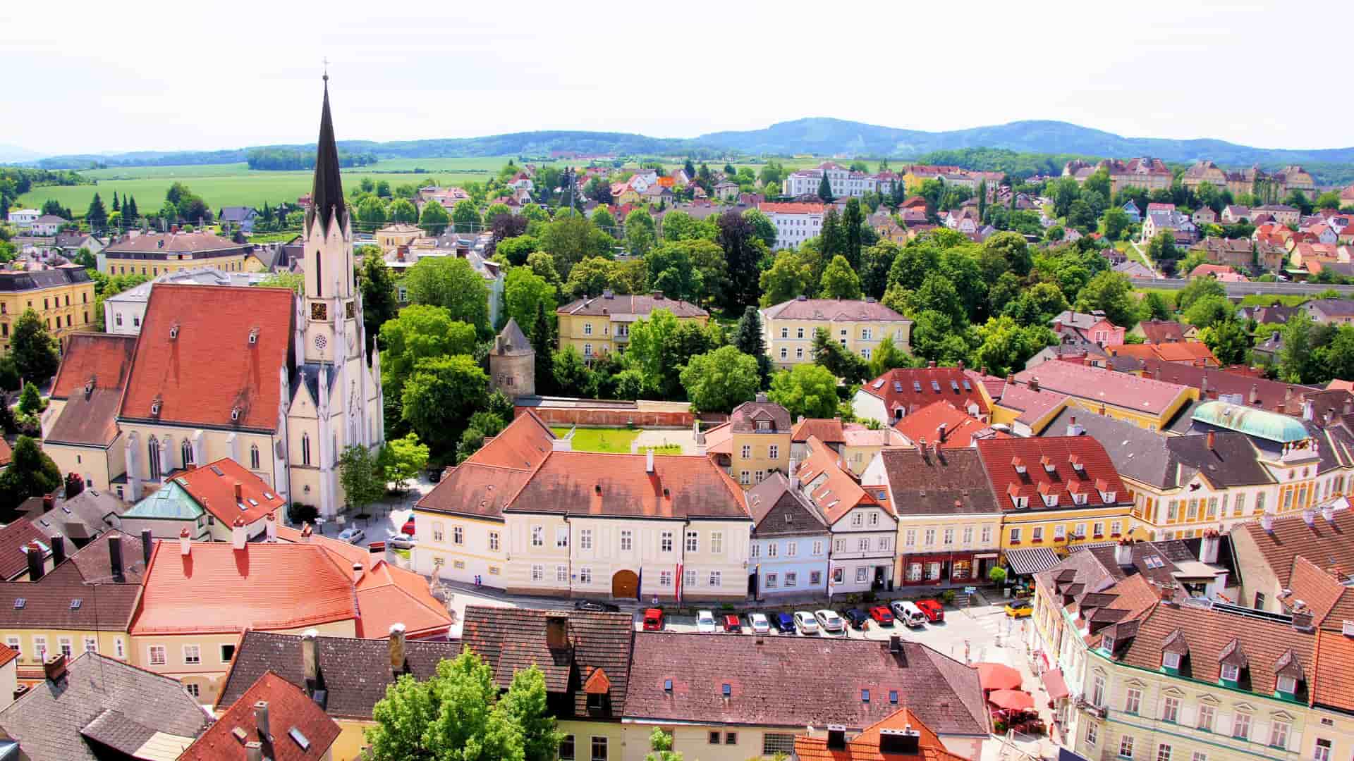 A scenic aerial view of the historic town of Melk, Austria, with a prominent church steeple and traditional red-roofed buildings surrounded by rolling green hills.