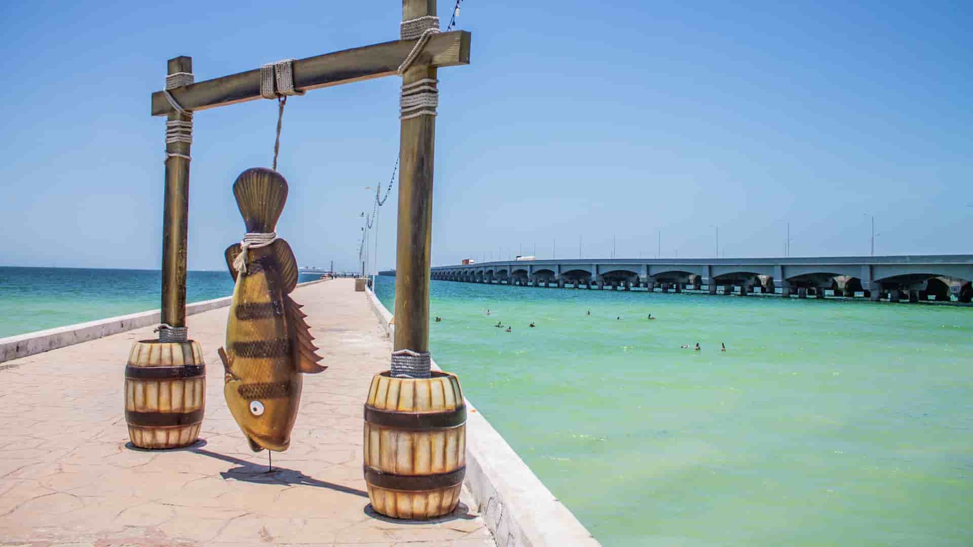 A vibrant shot of the long pier in Progreso, Mexico, with a large, decorative fish hanging from a wooden frame and the highway bridge visible in the distance over the turquoise sea.