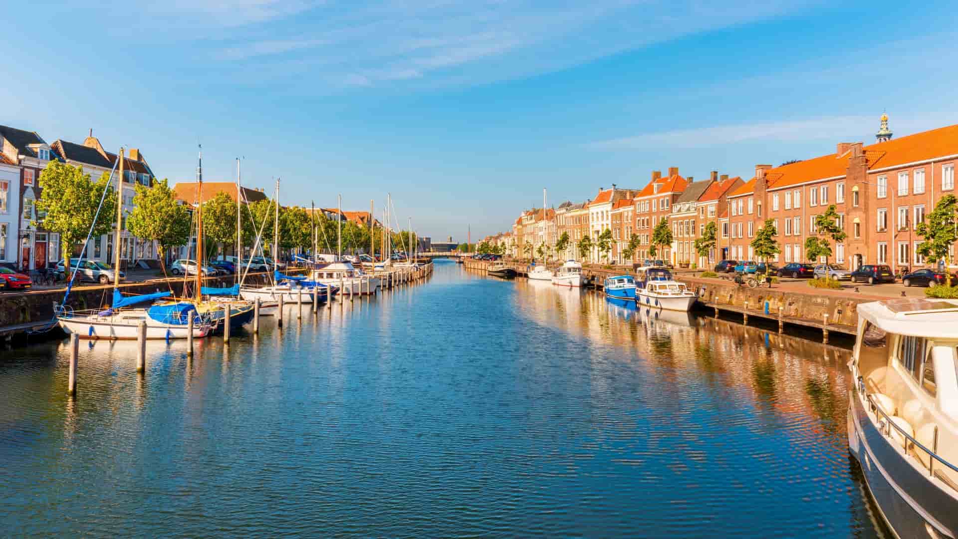 A scenic view of a tranquil canal in Middelburg, Netherlands, with traditional Dutch houses and moored boats lining the waterway under a clear blue sky.