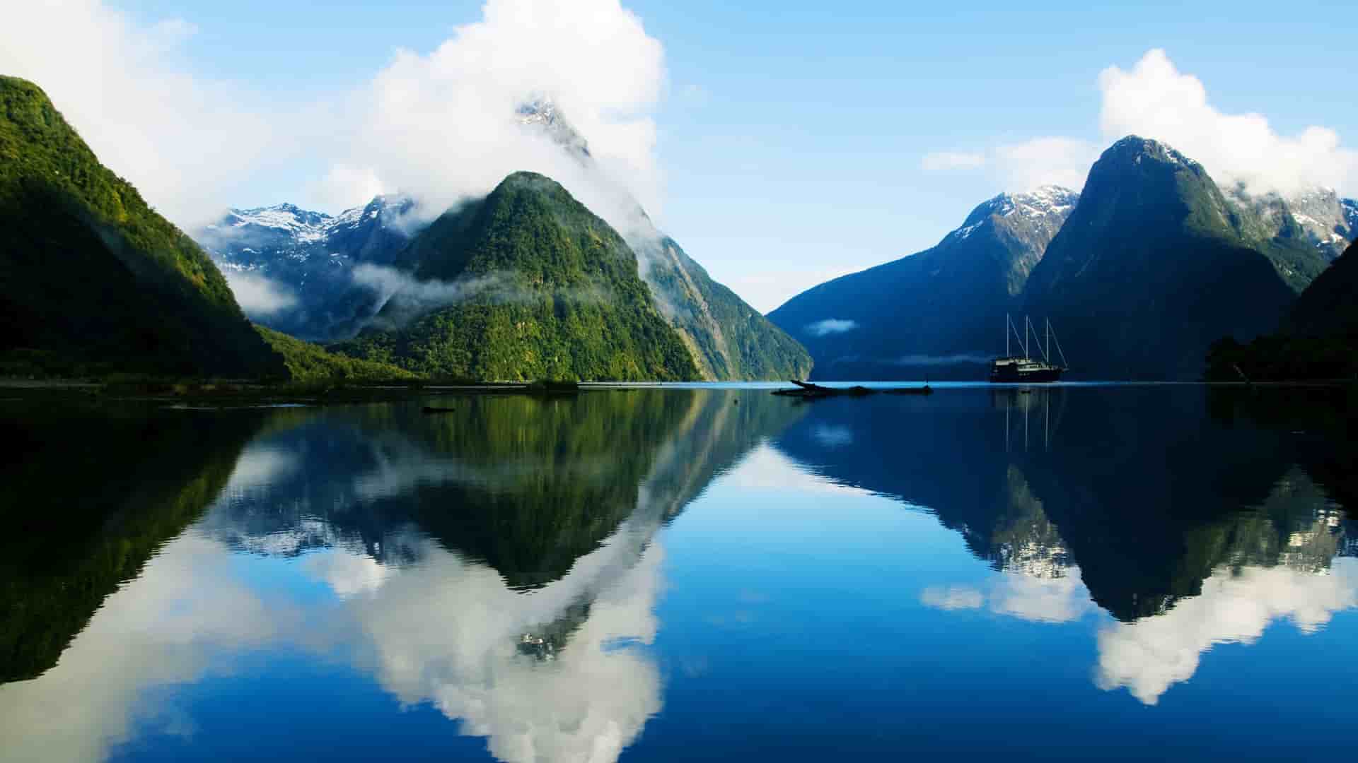A serene and majestic landscape of Milford Sound in New Zealand, with a large boat cruising on the still water. The scene features stunning reflections of the snow-capped mountains and clouds on the water.