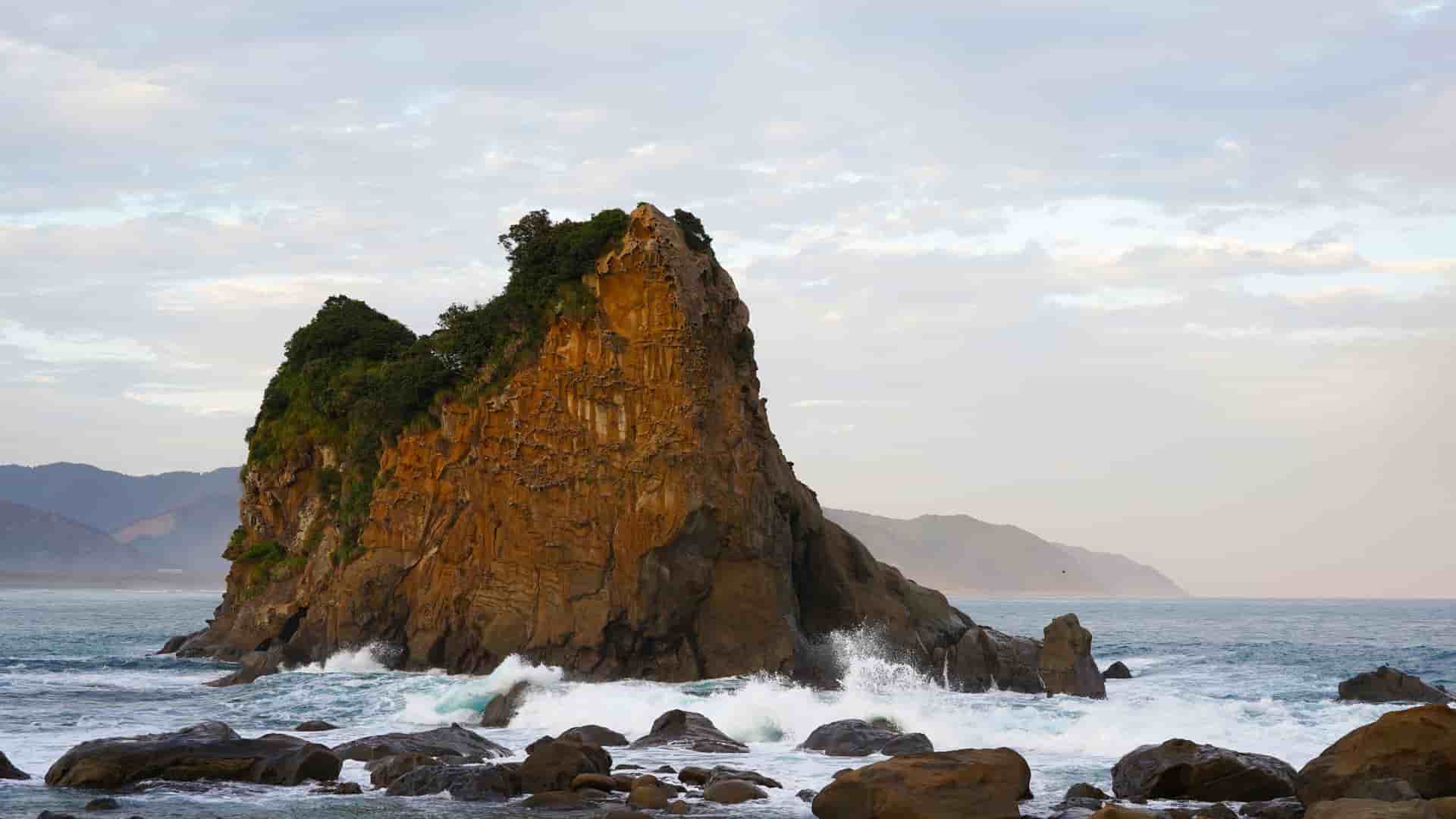 A massive, rugged sea stack with trees growing on its top, located in the coastal waters of Aburatsu, Miyazaki, Japan, with waves crashing against its rocky base.