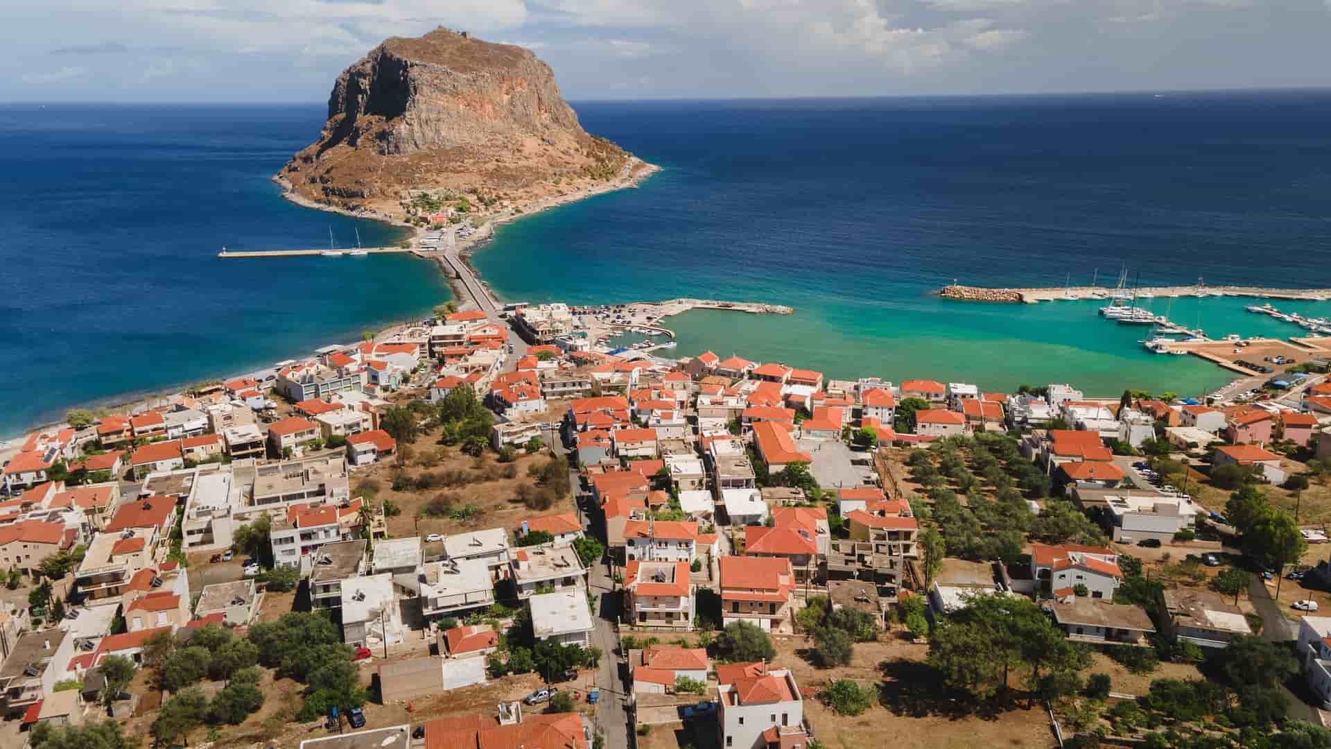 An aerial view of the historic town of Monemvasia, Greece, with its red-roofed buildings nestled on a small peninsula connected to the mainland by a single causeway.