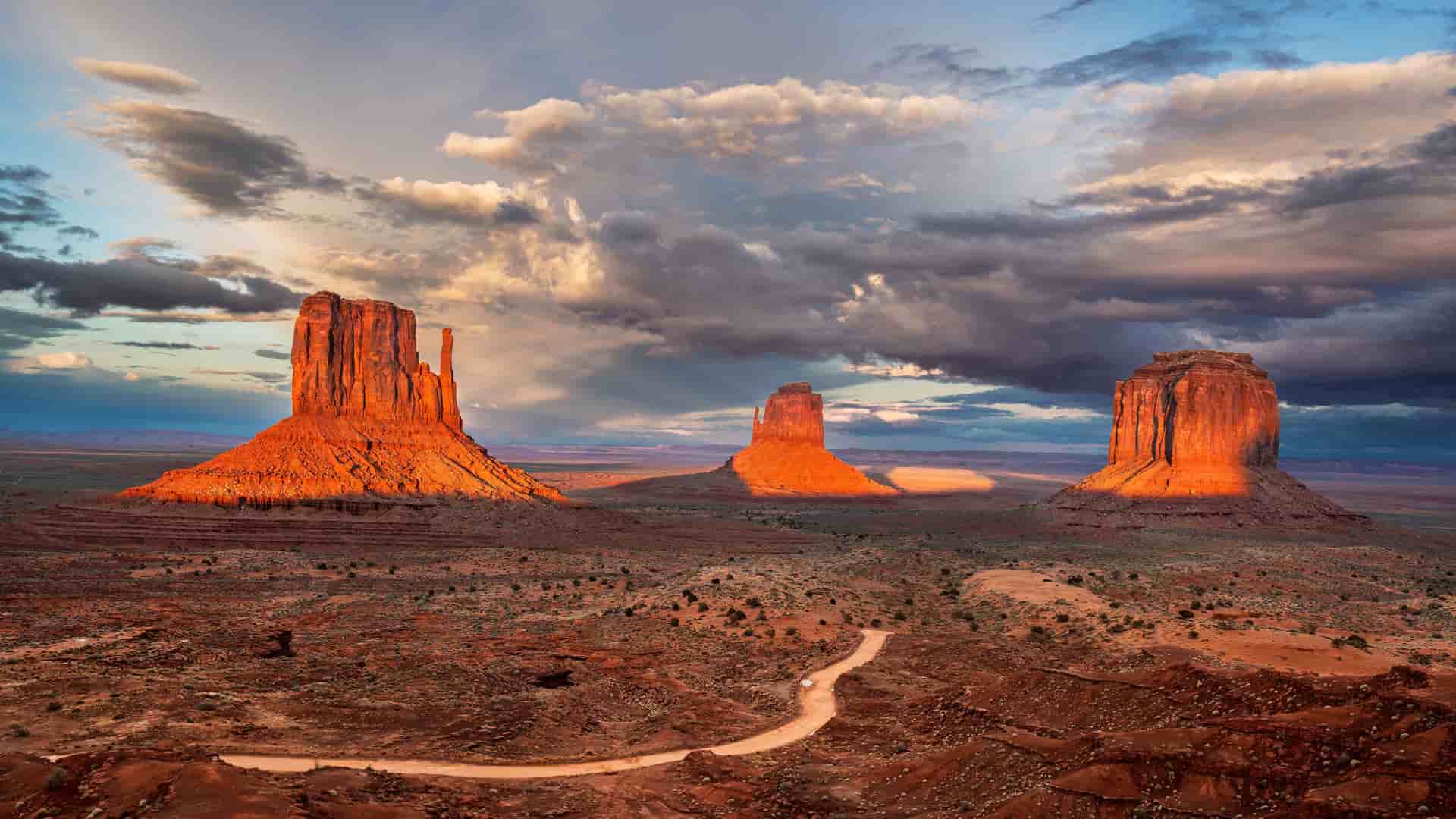 "A dramatic panoramic view of the iconic sandstone buttes and mesas in Monument Valley, Utah, glowing orange at sunset under a moody, cloudy sky.  "