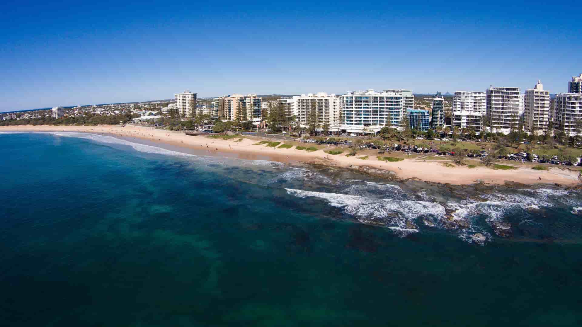An aerial view of the sunny Mooloolaba Beach, Australia, showcasing a long stretch of golden sand, a calm blue ocean, and a line of tall resort buildings on the coastline.