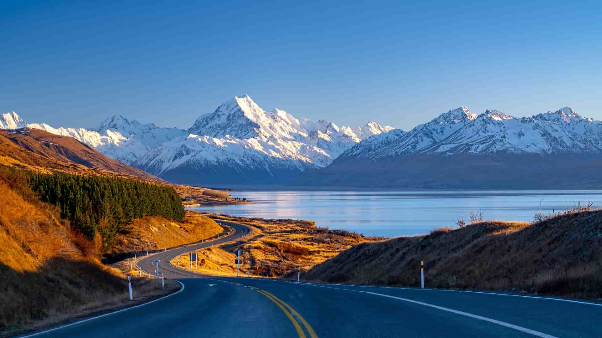 "A beautiful panoramic shot of Mount Cook National Park in New Zealand, with the iconic snow-capped Aoraki Mount Cook mountain range overlooking the bright blue waters of Lake Pukaki at sunrise.  "