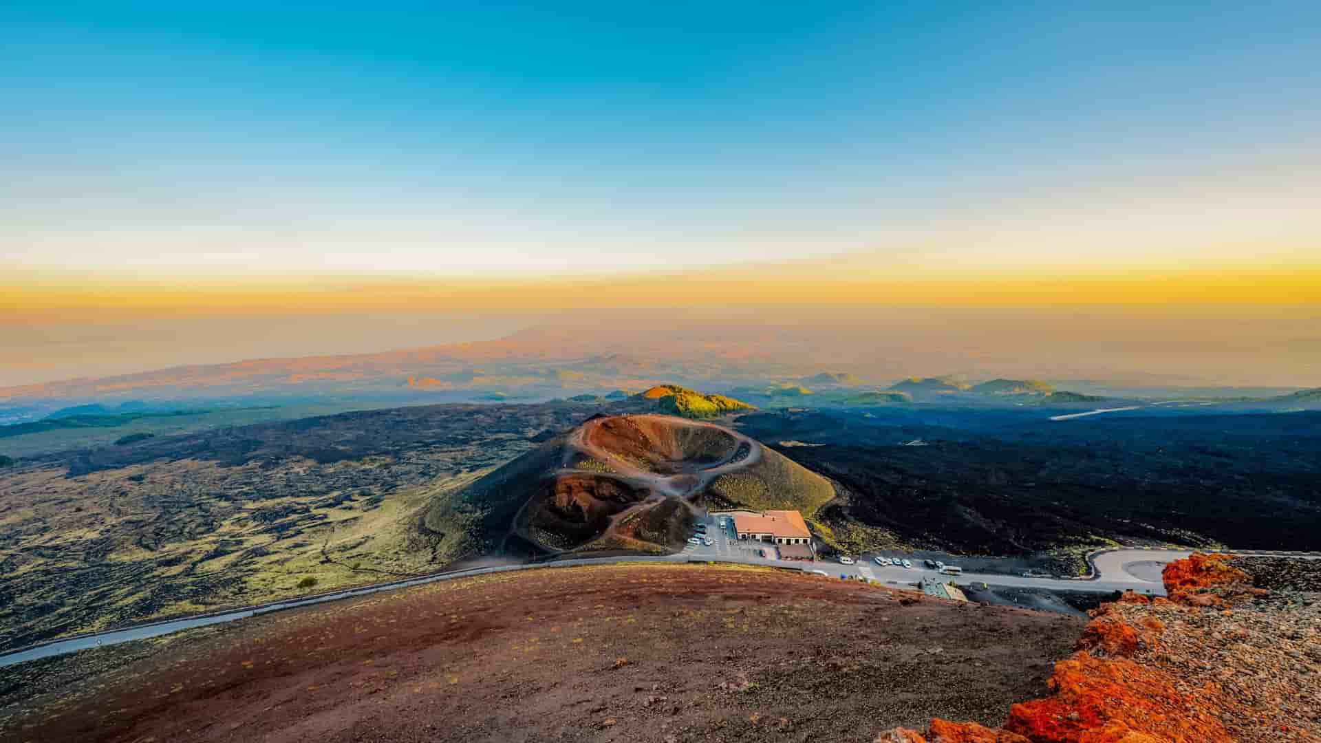 A high-angle view of Mount Etna, Italy, shows a visitor center on a road, surrounded by dark volcanic ash and rock, at sunset.