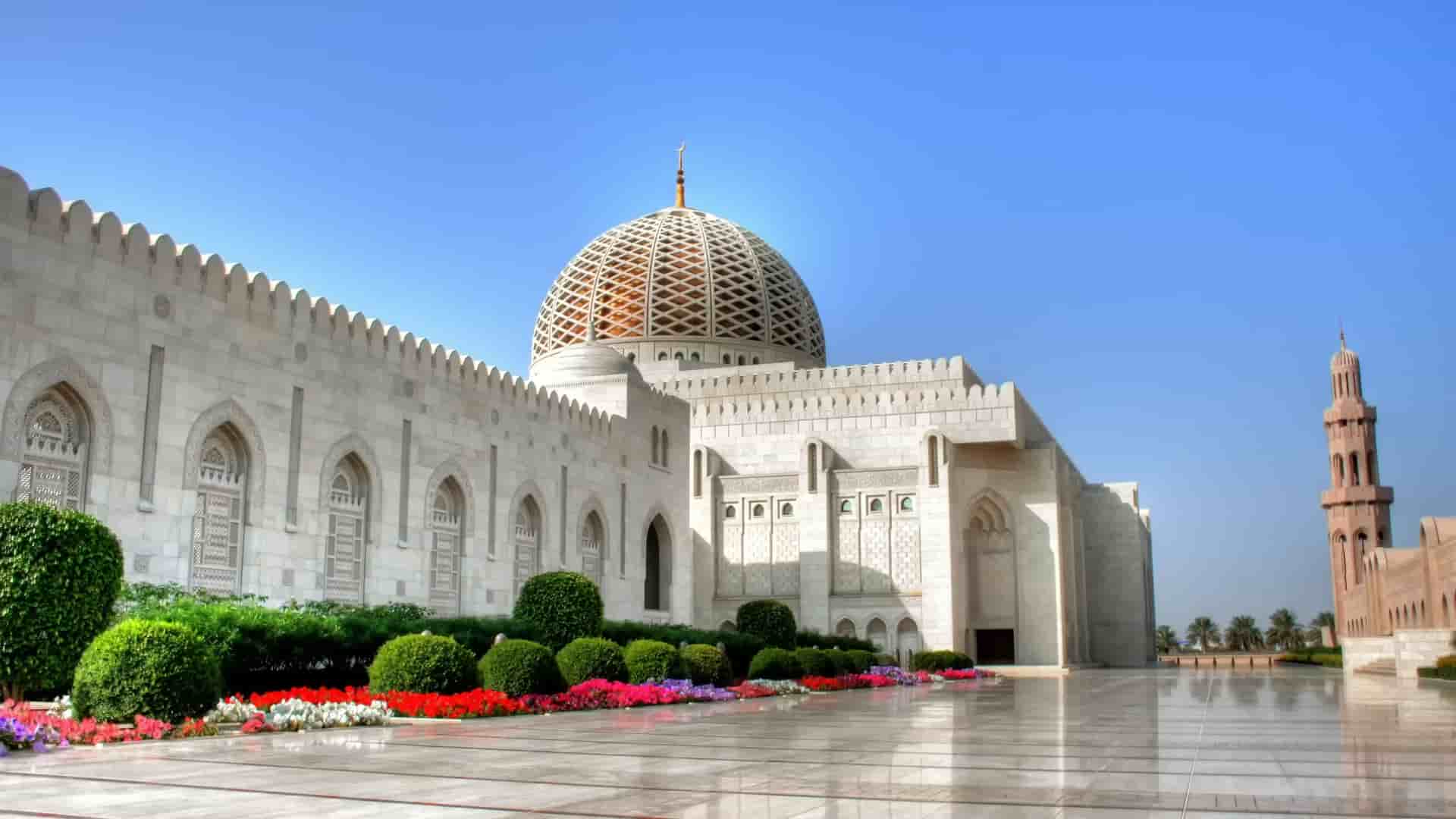 A side view of the grand Sultan Qaboos Grand Mosque in Muscat, Oman, featuring its majestic dome and minaret under a clear blue sky.