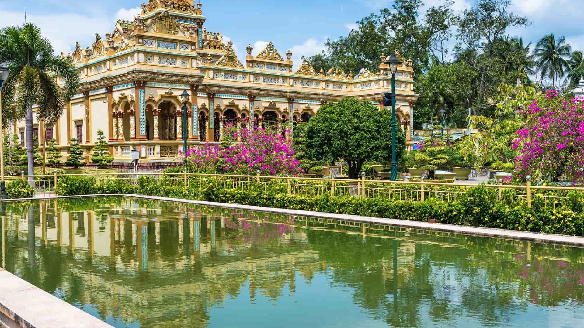 A colorful, ornate temple in My Tho, Vietnam, with intricate architectural details, surrounded by lush gardens, palm trees, and a tranquil reflecting pond.