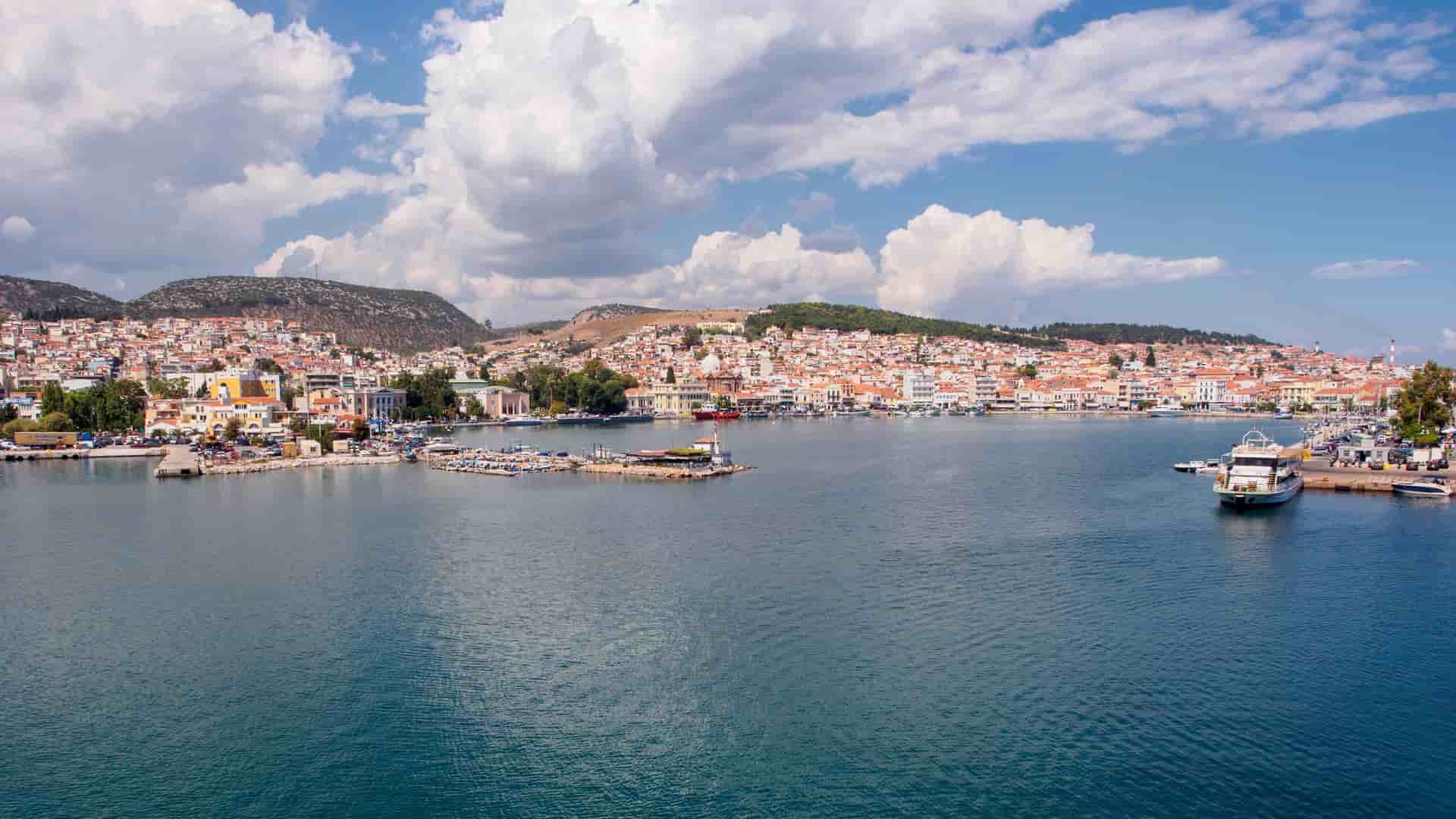 A broad view of the city of Mytilene on the island of Lesbos, with buildings and a harbor lining the waterfront and lush hills in the background.