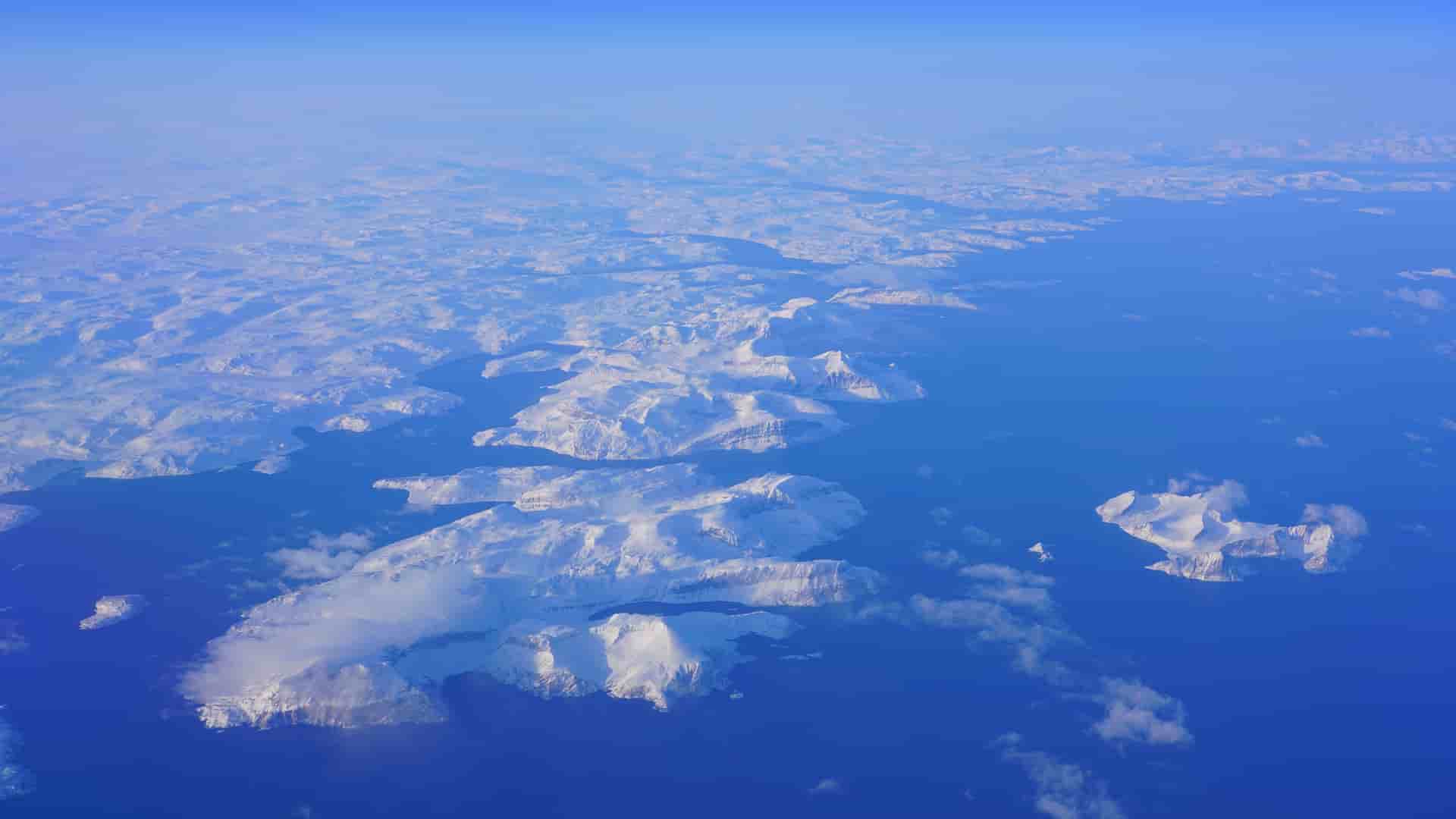An aerial view of the snow-covered mountains and islands along the coast of Nain, Canada, with vast blue water and a distant shoreline under a clear sky.