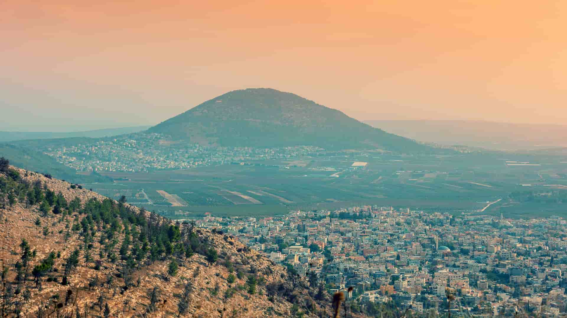 "A high-angle view of the city of Nazareth, Israel, nestled in a valley with Mount Tabor and the surrounding landscape in the distance at sunset.  "